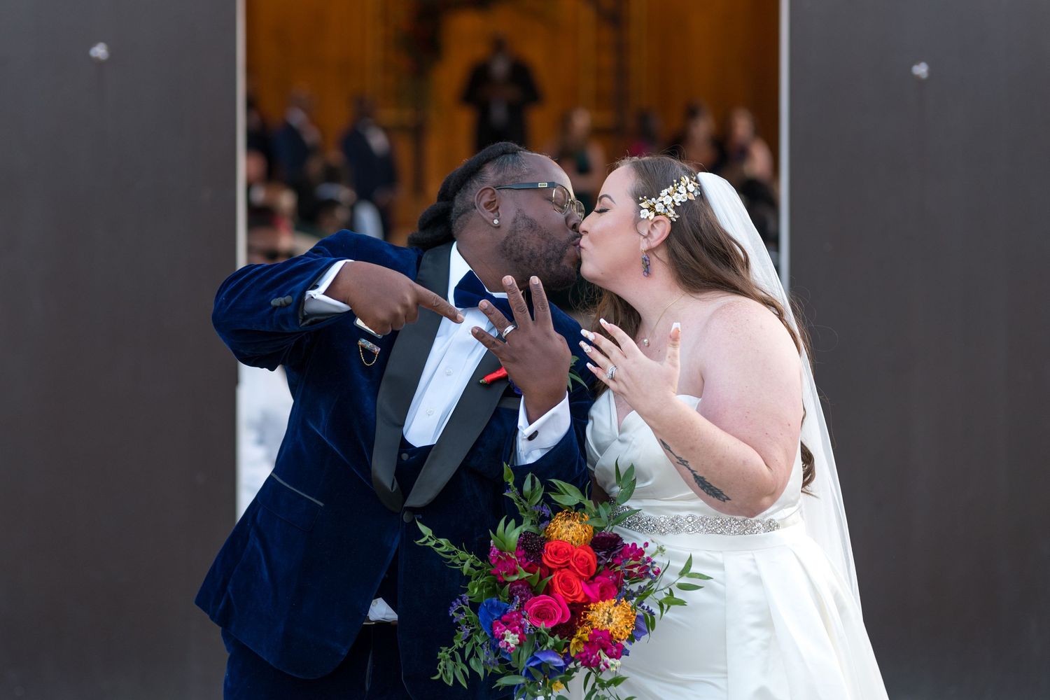 Newlywed couple sharing a kiss at the altar, bride holding colorful bouquet, groom in navy suit, both showing wedding rings.
