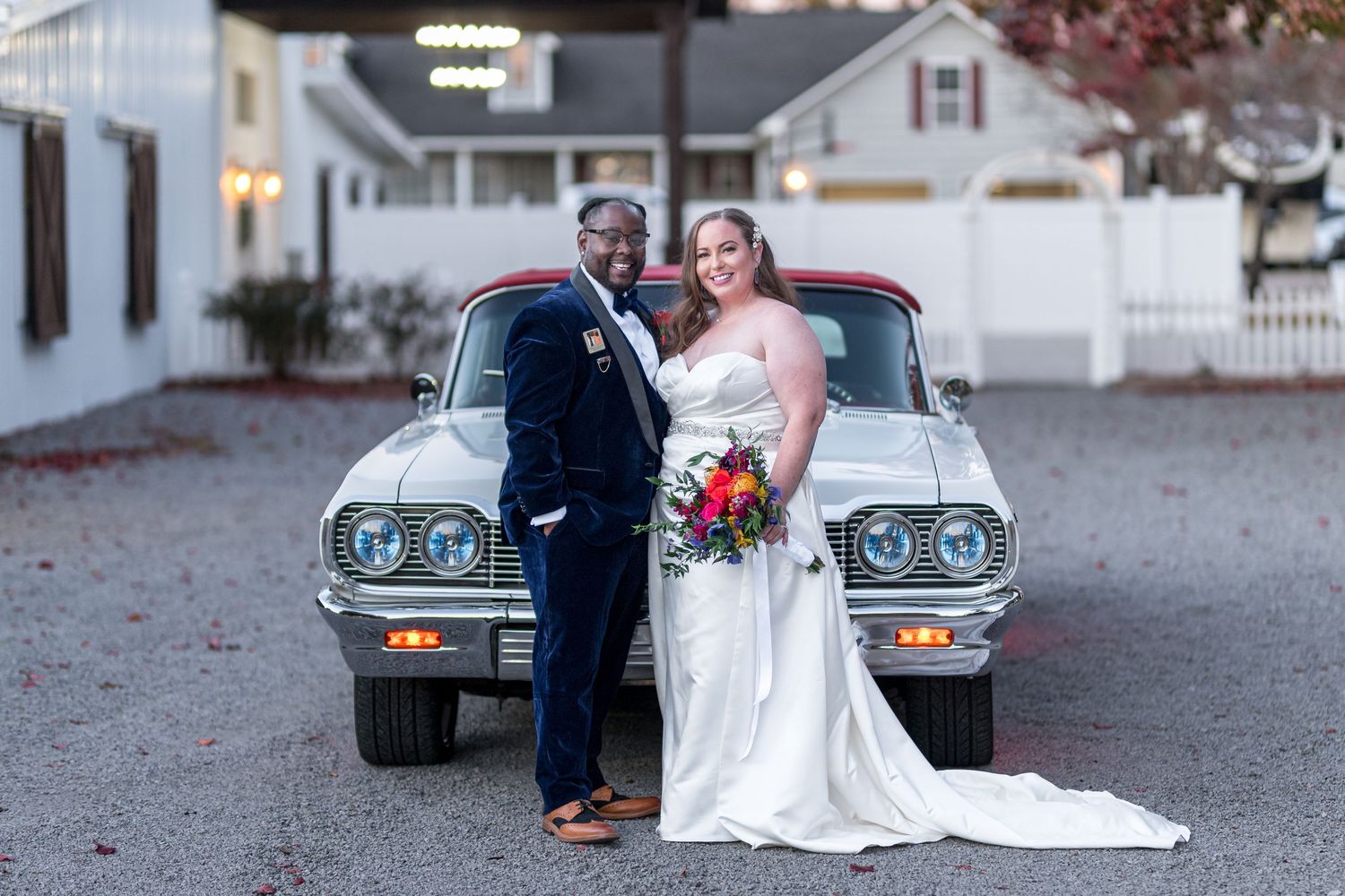 Bride and groom posing together in front of a vintage classic car at an outdoor wedding venue.