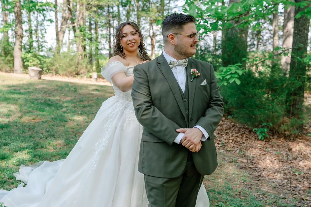 Bride in white ball gown and groom in olive suit pose together outdoors in a lush green forest setting.