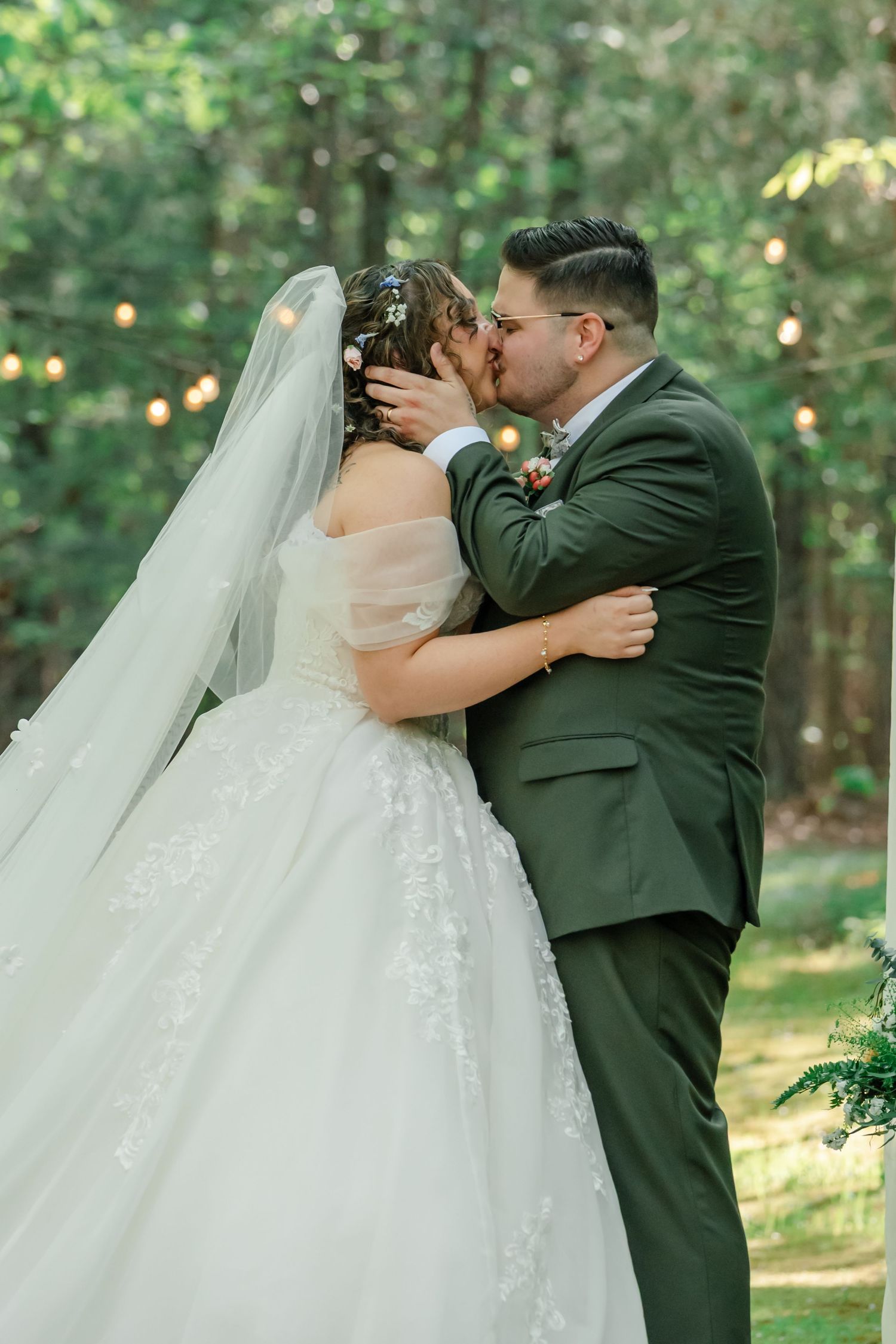 Bride and groom share first kiss outdoors in lush green forest setting, bride in lace ballgown with veil, groom in olive suit.