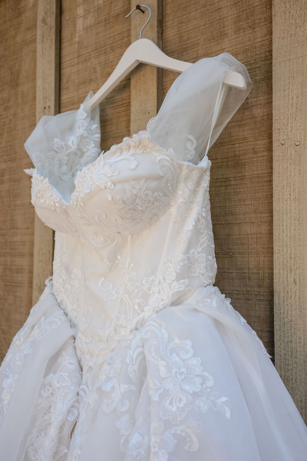 Elegant white lace ball gown wedding dress hanging on a wooden wall, featuring intricate floral embroidery and sheer straps.