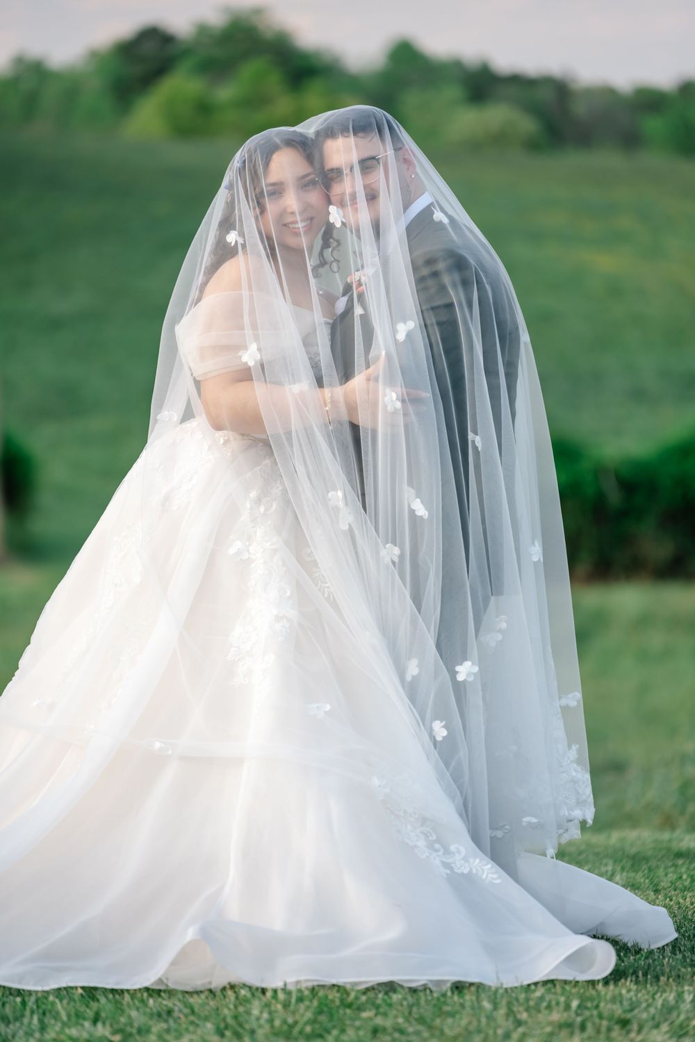 Bride and groom share a romantic moment under a floral veil in a lush green outdoor setting.