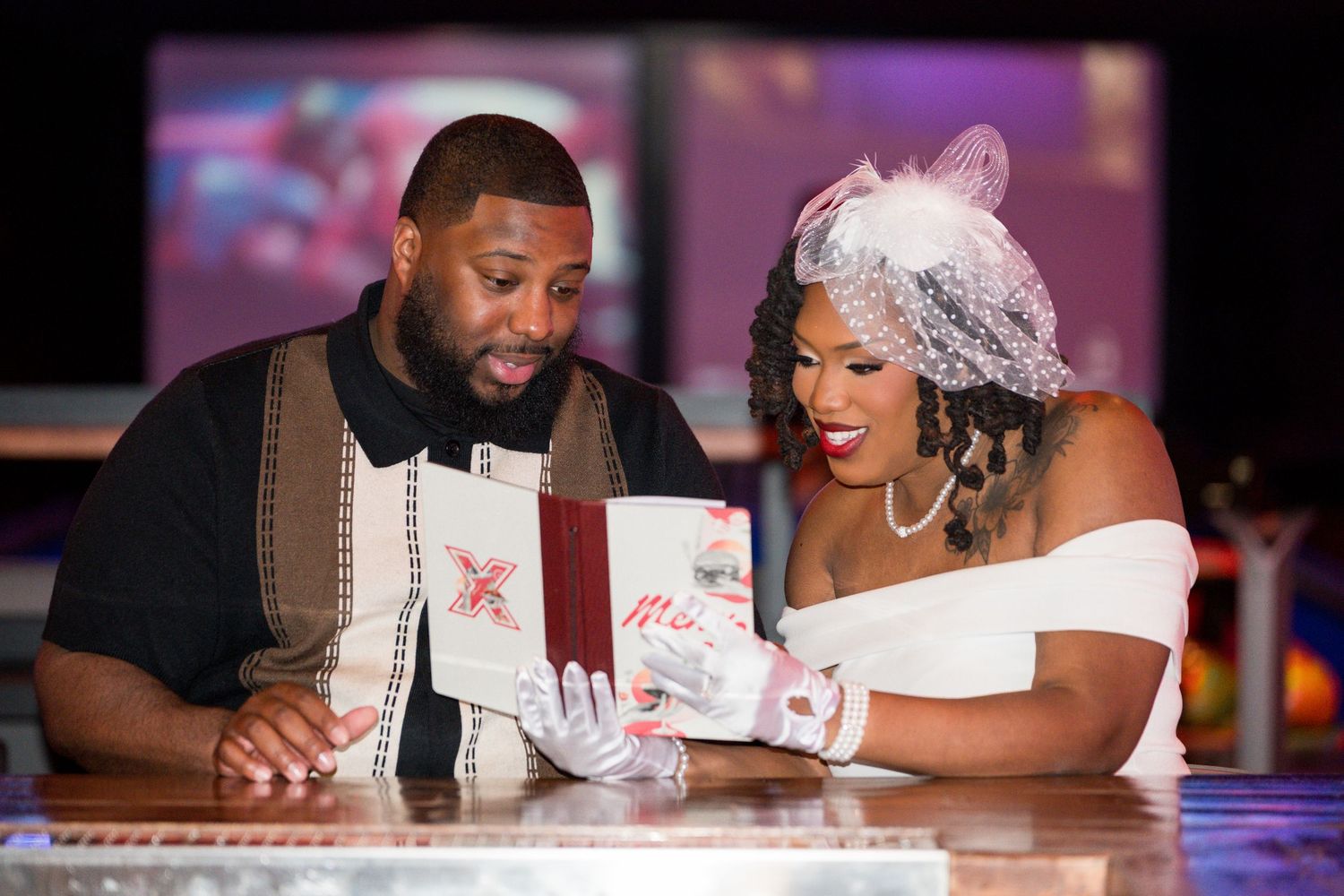 Couple in wedding attire reviewing a decorative book at a bar, woman in white dress with veil, man in suspenders.