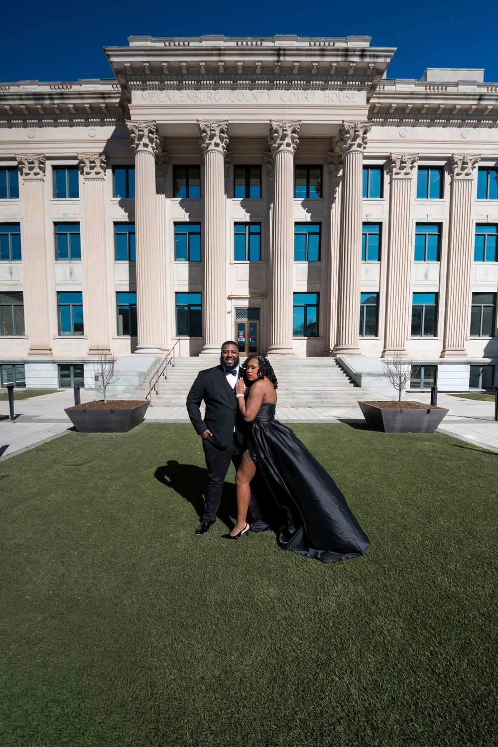 Elegant couple in formal black attire posing in front of a grand neoclassical building with tall columns on a sunny day.