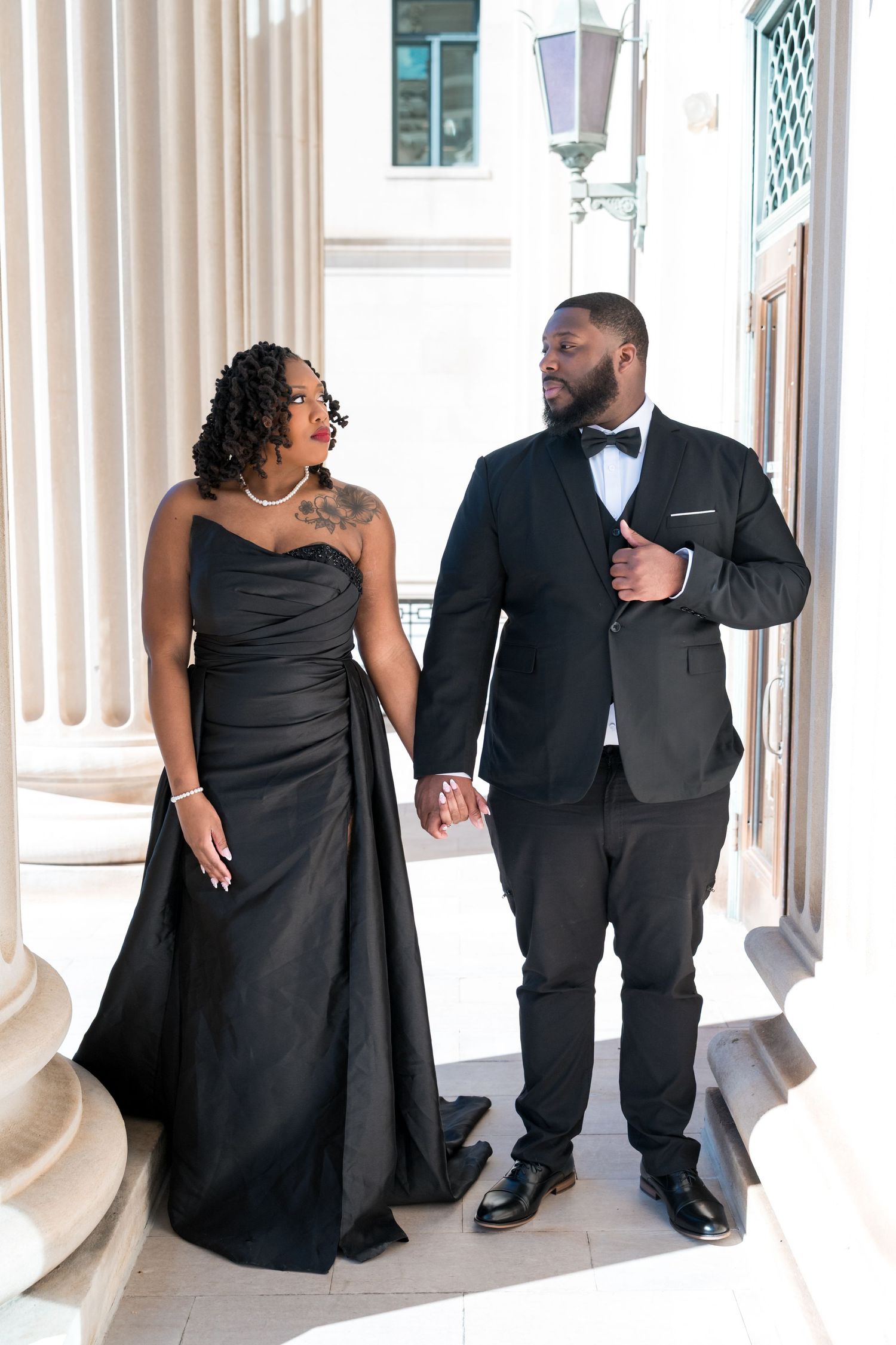 Elegant couple in matching black formal attire holding hands beside white columns at an upscale venue.
