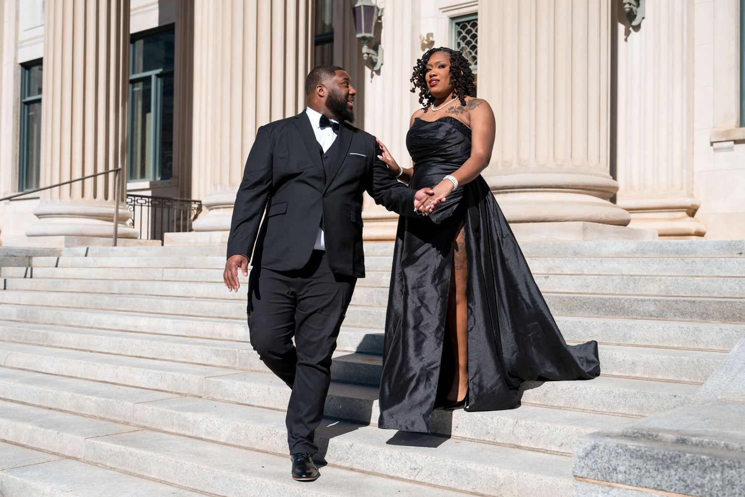 Elegant couple in matching black formal attire descending grand stone steps with white columns.