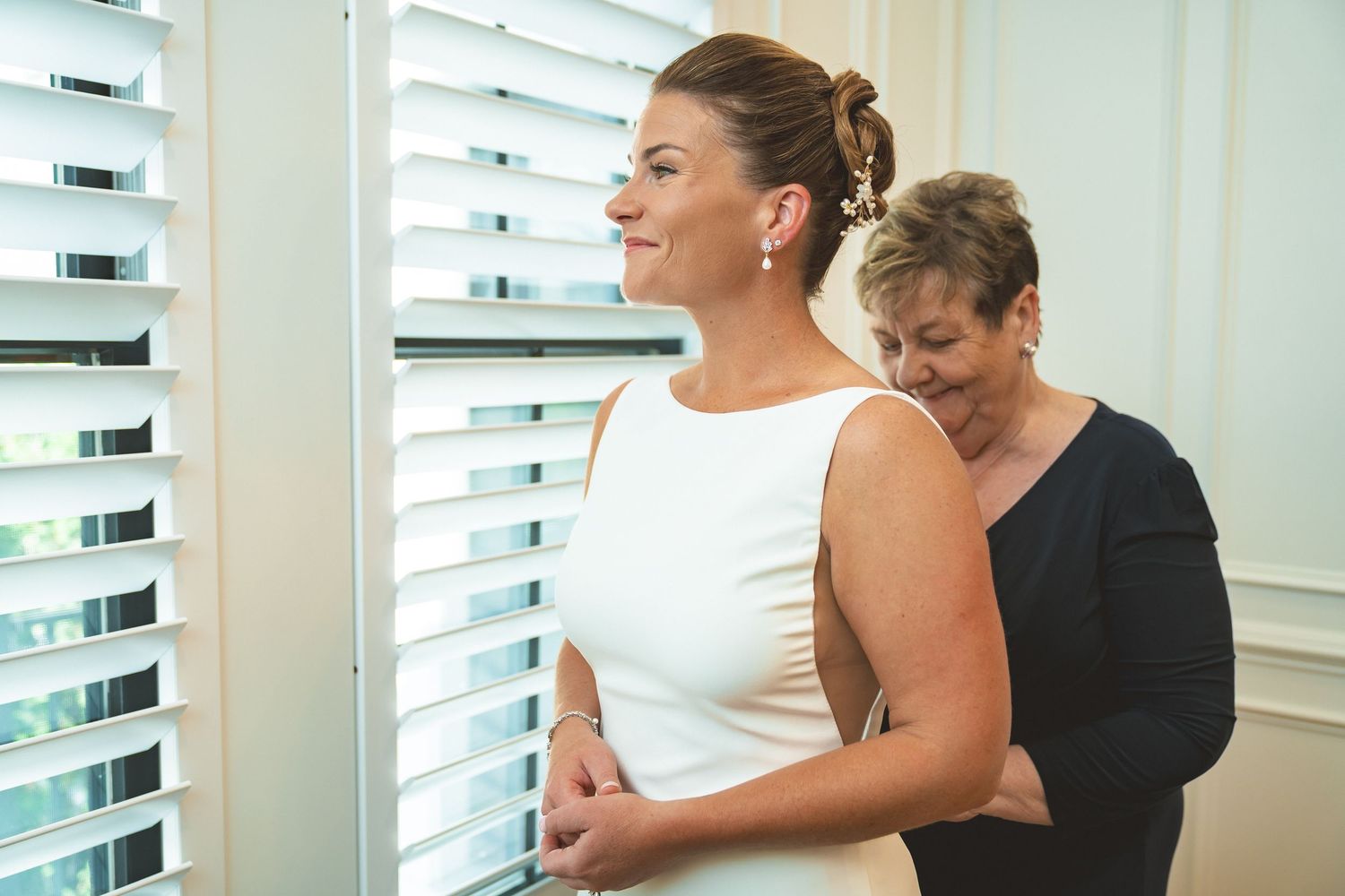 Bride in white dress stands by window blinds as older woman helps her prepare for wedding day.