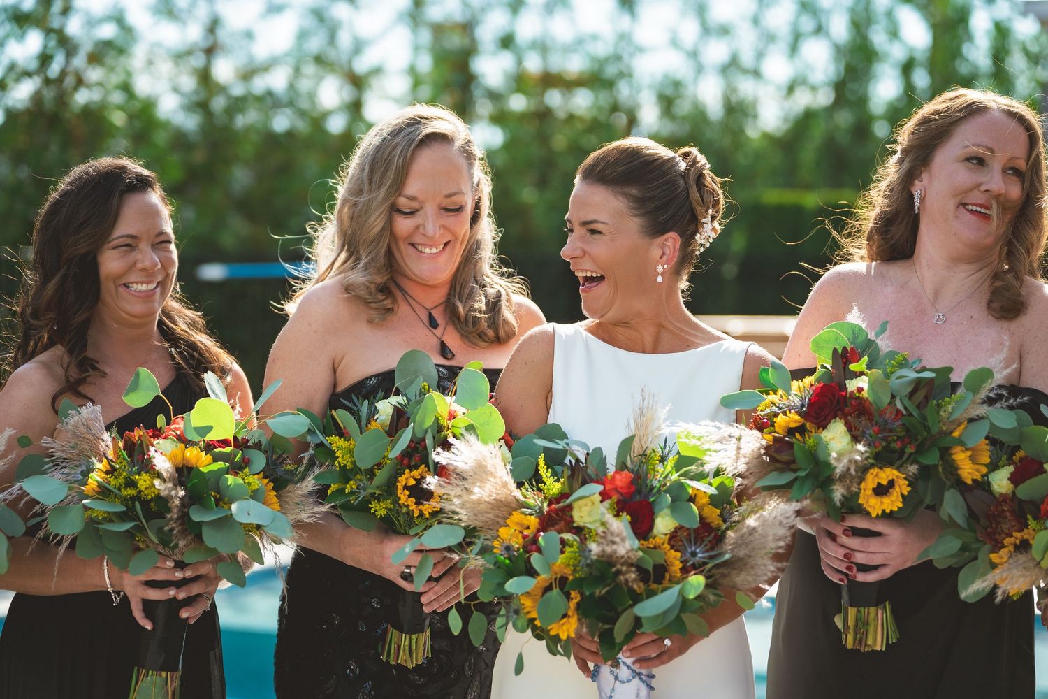 Smiling bride with bridesmaids in dark dresses holding vibrant sunflower and greenery bouquets outdoors.