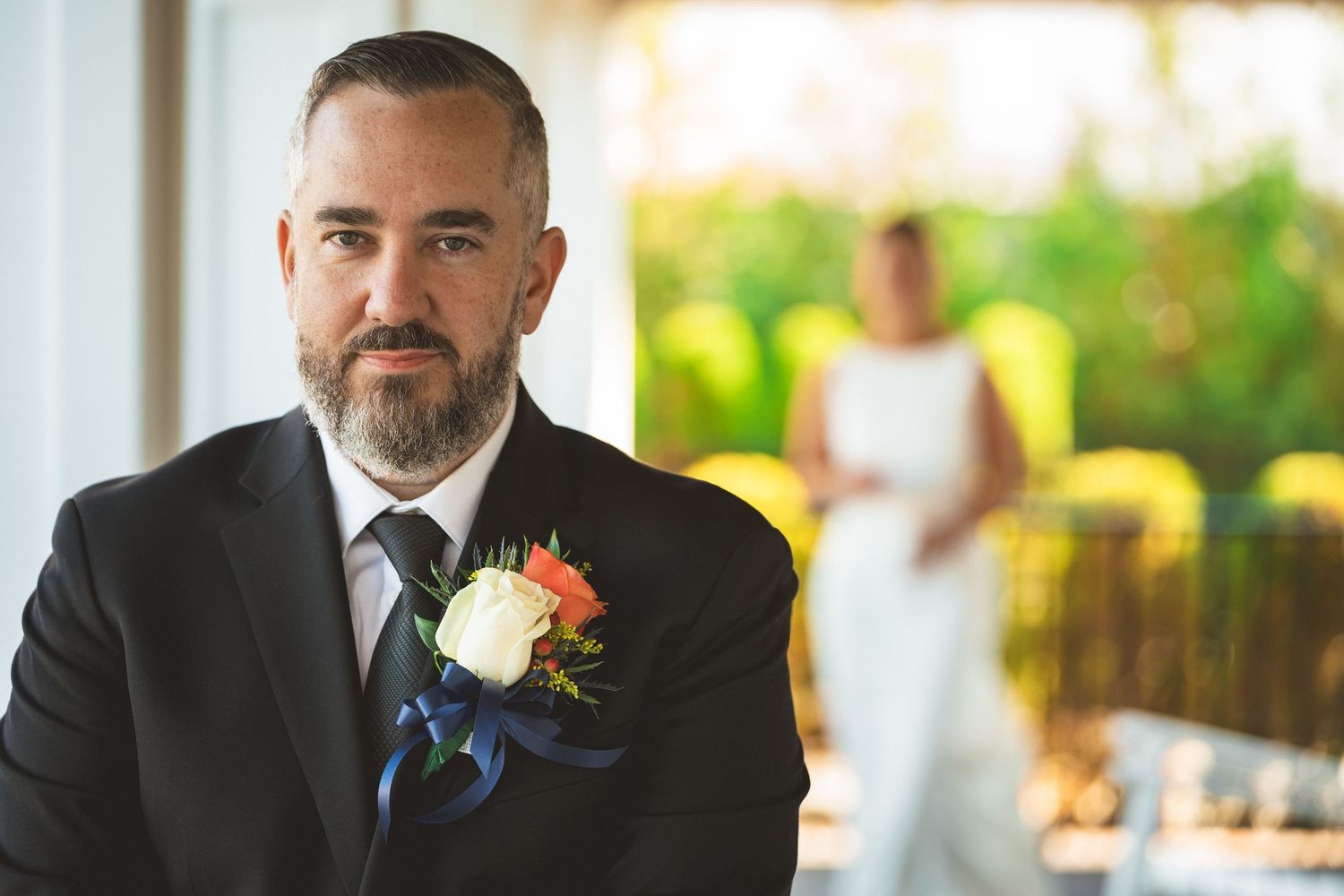 Groom in black suit with floral boutonniere stands in foreground, bride in white dress visible in background.