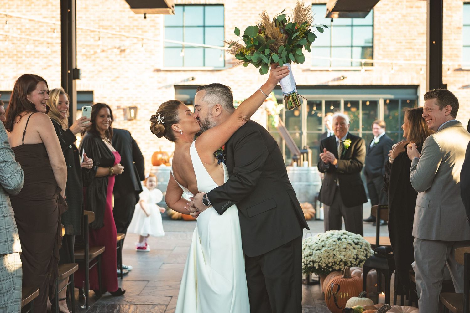 Newlywed couple sharing a kiss at wedding ceremony, bride raising bouquet, guests applauding in elegant brick venue.