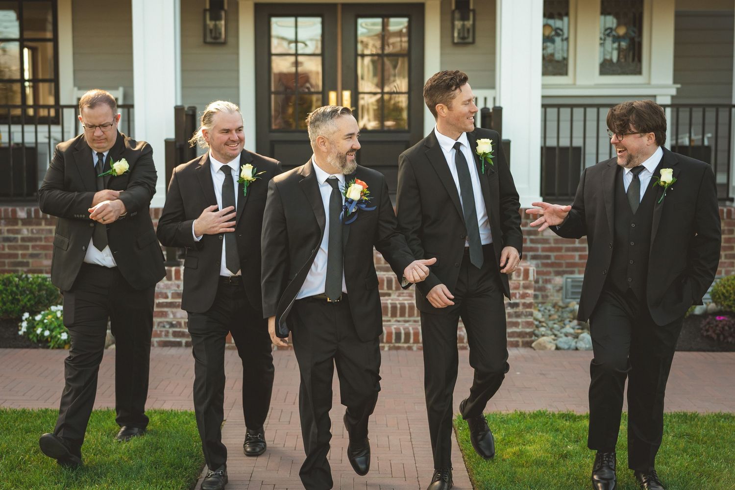 Five groomsmen in black suits with boutonnieres walk and laugh together outside an elegant venue.