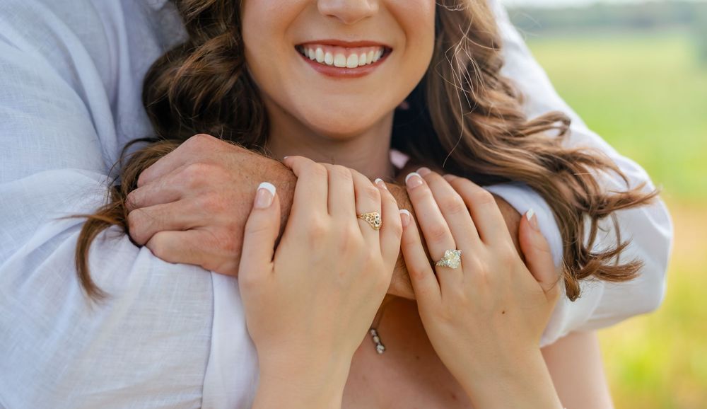 Close-up of smiling bride wearing engagement ring, embraced by groom in white shirt outdoors.