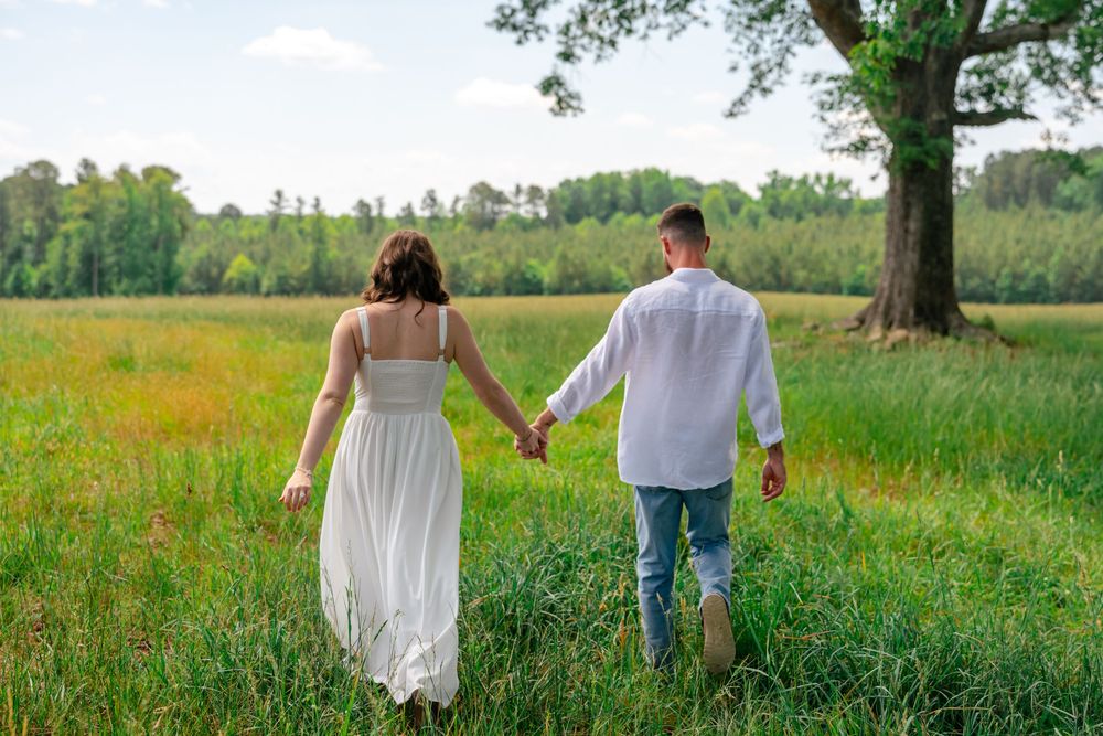 Couple holding hands walking through a lush green meadow with wildflowers and trees in the background.