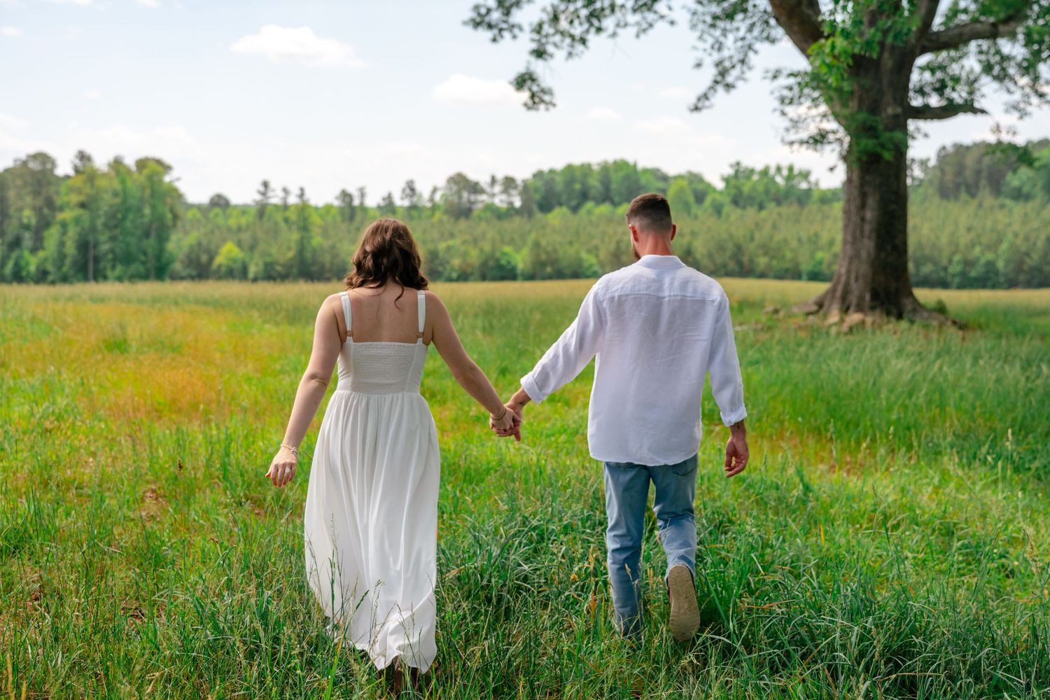Couple holding hands walking through a lush green meadow with wildflowers and trees in the background.