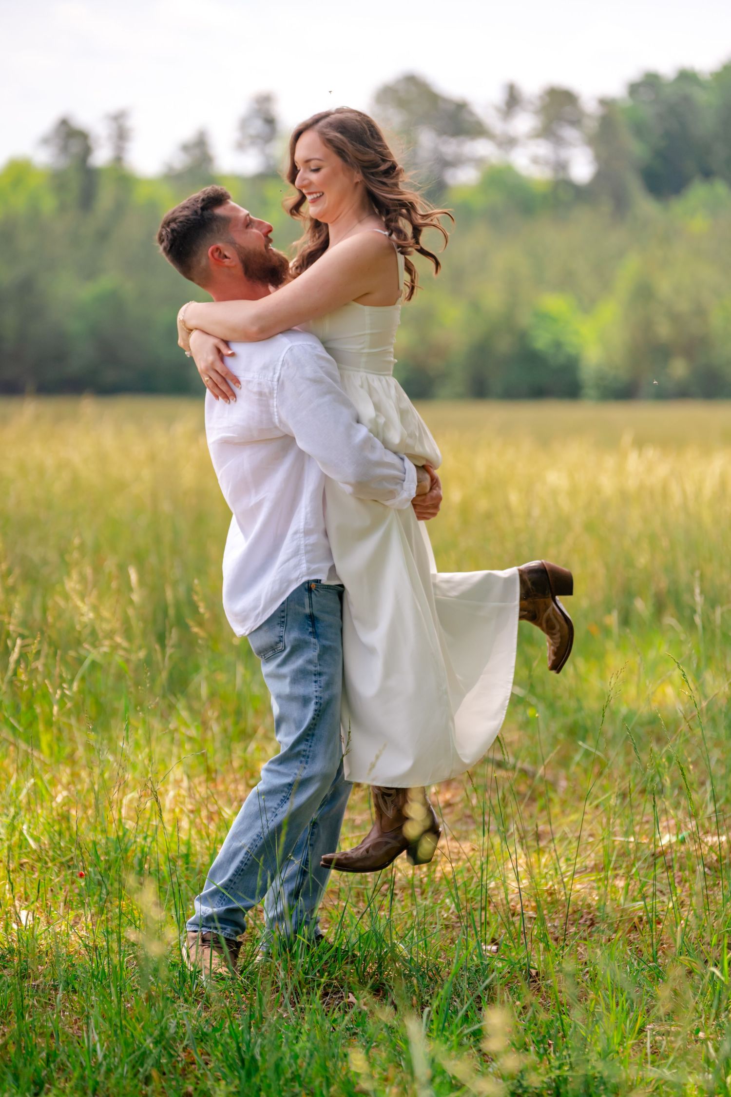 A groom lifts his smiling bride in a white dress and cowboy boots in a lush green field.