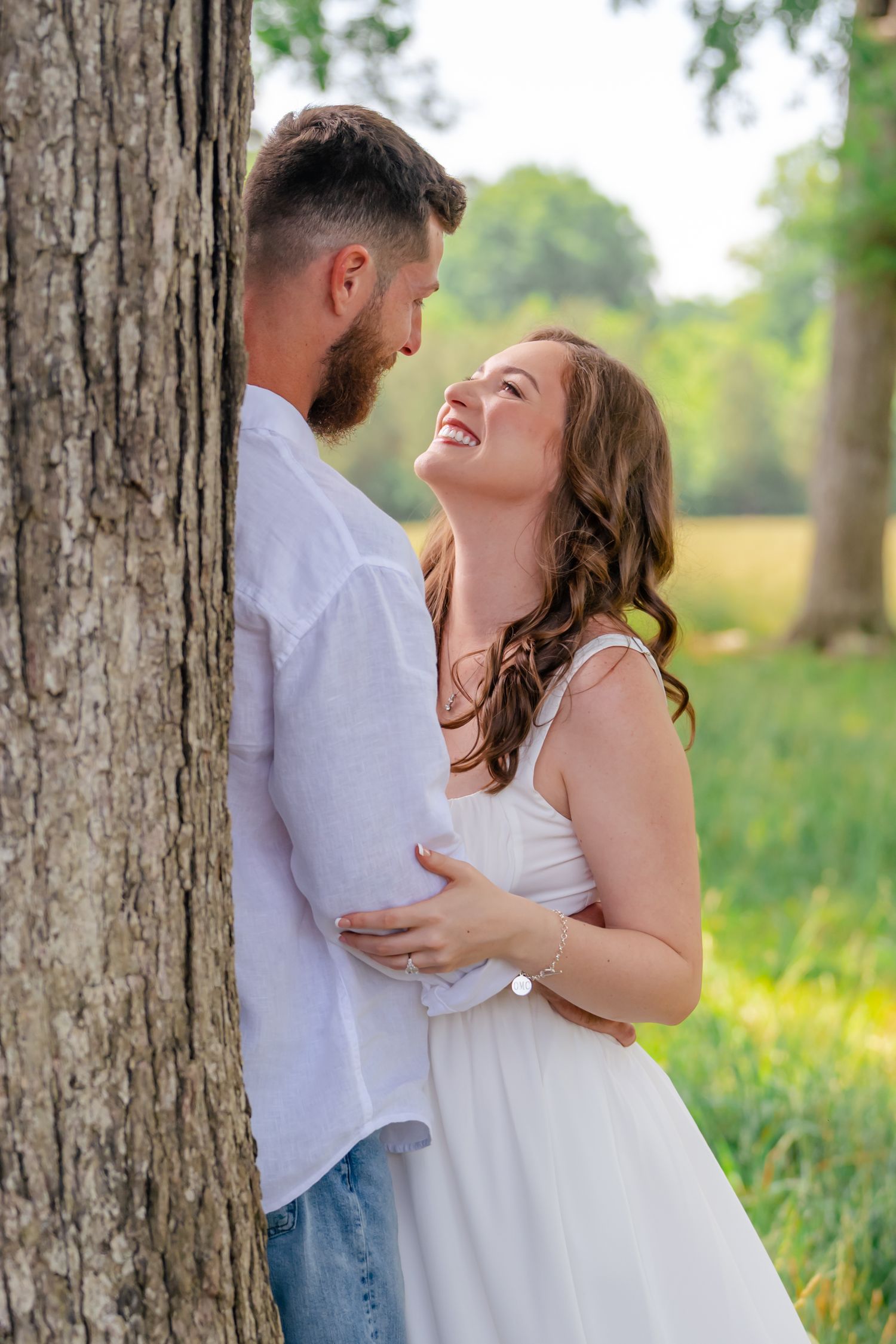 Happy couple embracing by a tree in a sunny park, woman in white dress smiling up at bearded man.
