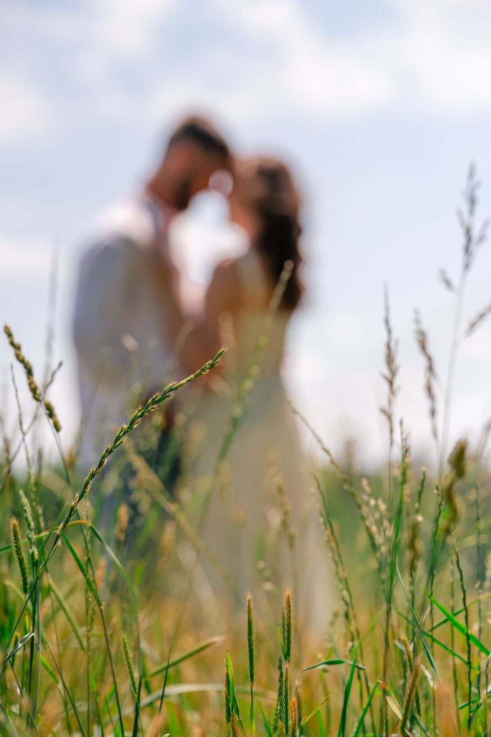 Couple sharing a romantic moment in a lush green field, with tall grass in the foreground and a blue sky behind.