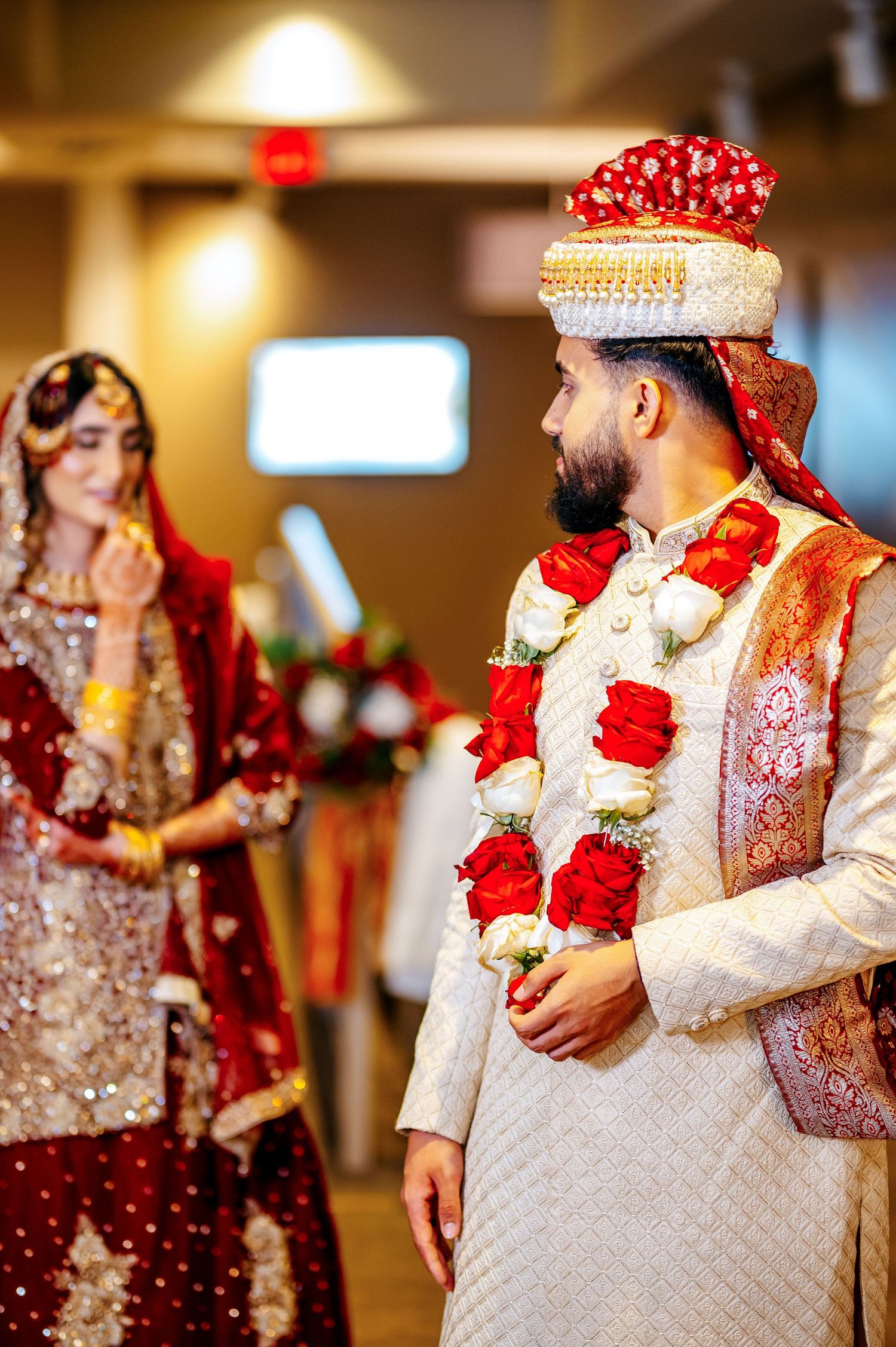 Groom in white sherwani with red floral garland stands beside bride in red bridal lehenga during Indian wedding ceremony.
