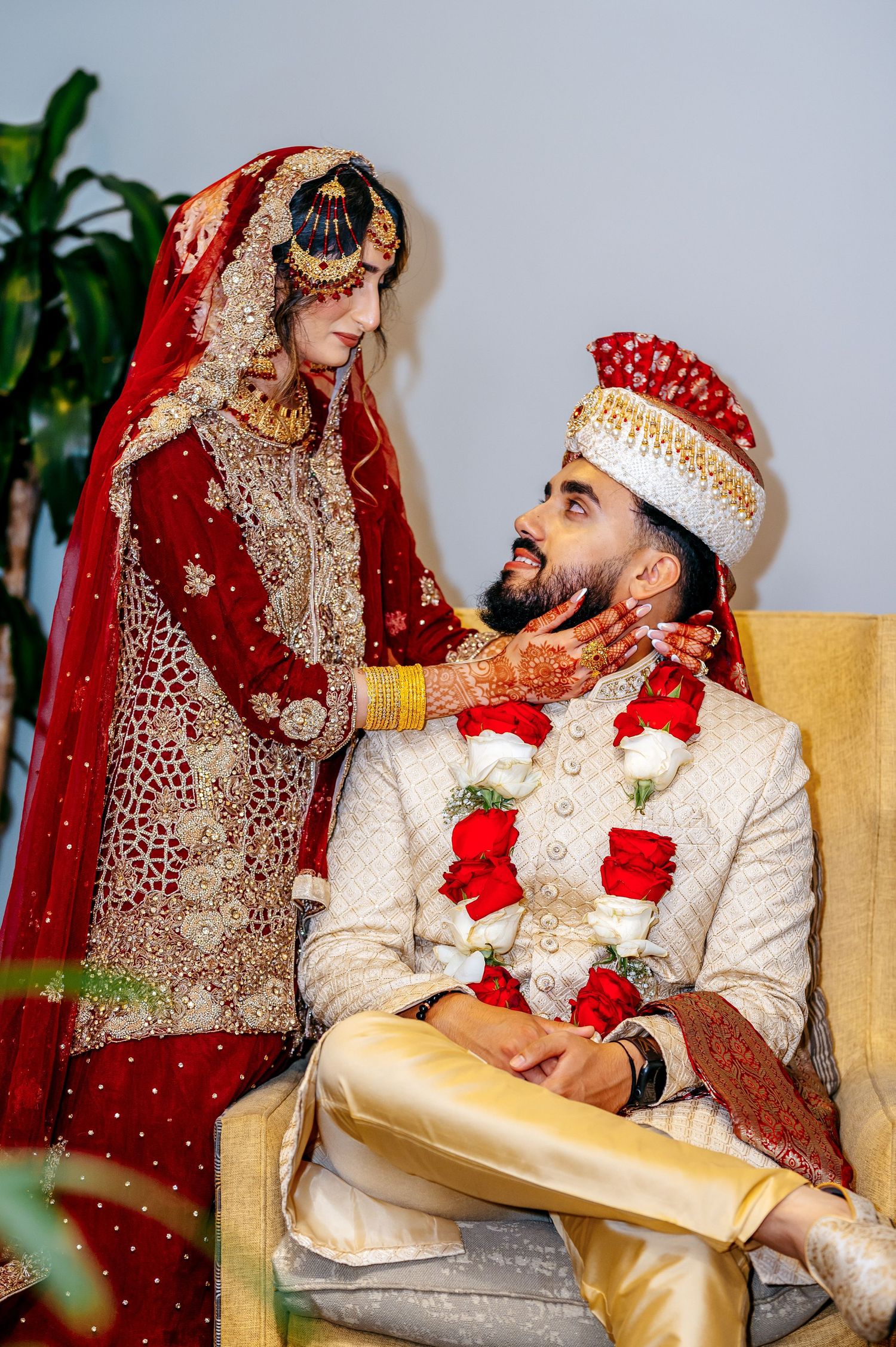 A bride in red bridal lehenga and groom in white sherwani share a tender moment during their South Asian wedding ceremony.