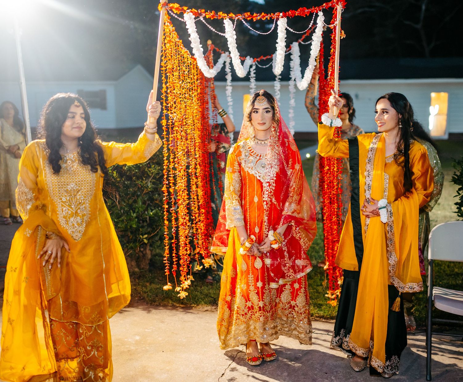 South Asian bride in red lehenga walks under floral canopy held by bridesmaids in yellow suits.