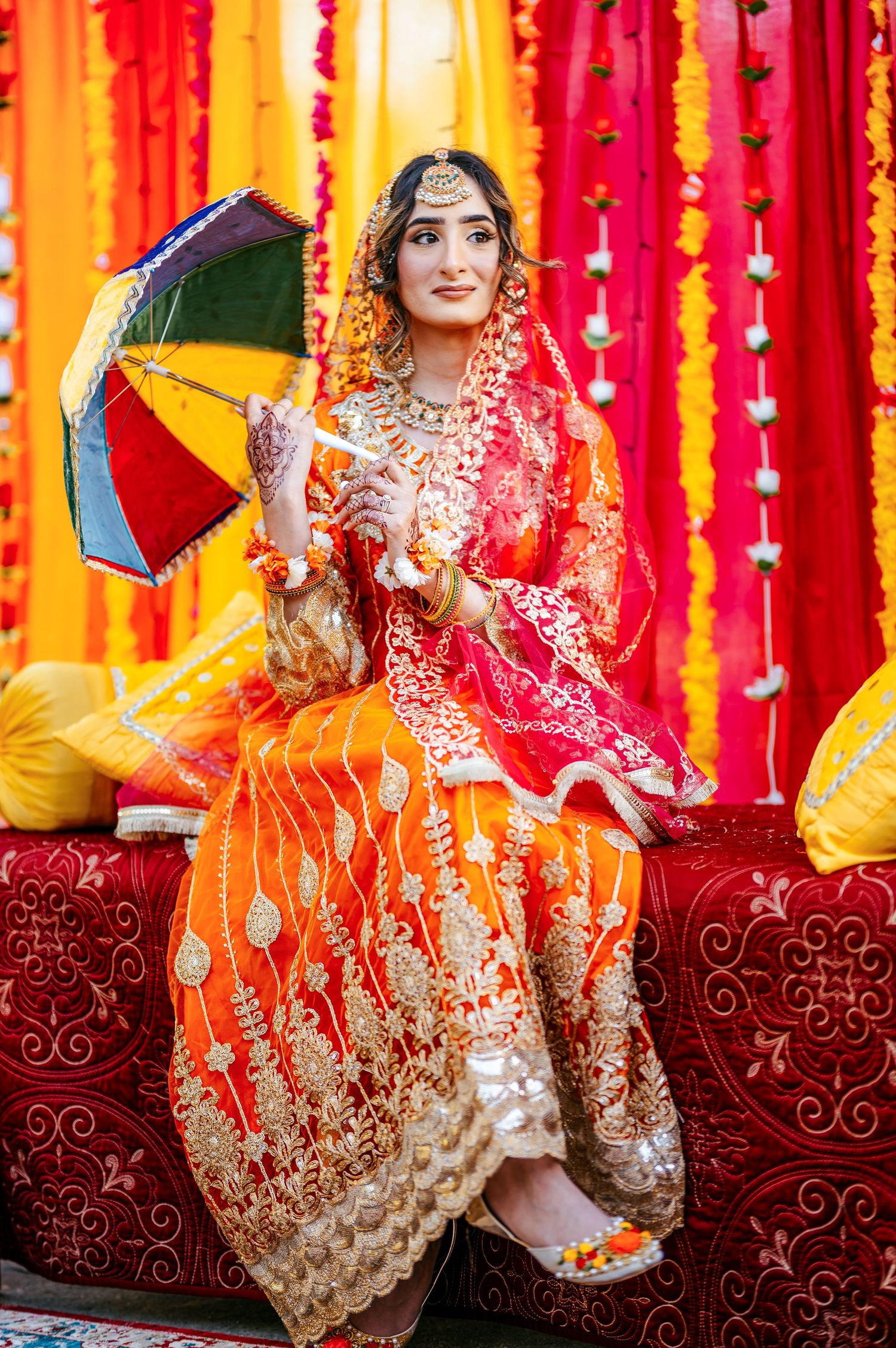 A bride in orange and red lehenga holds a colorful umbrella, seated on red sofa with floral backdrop.