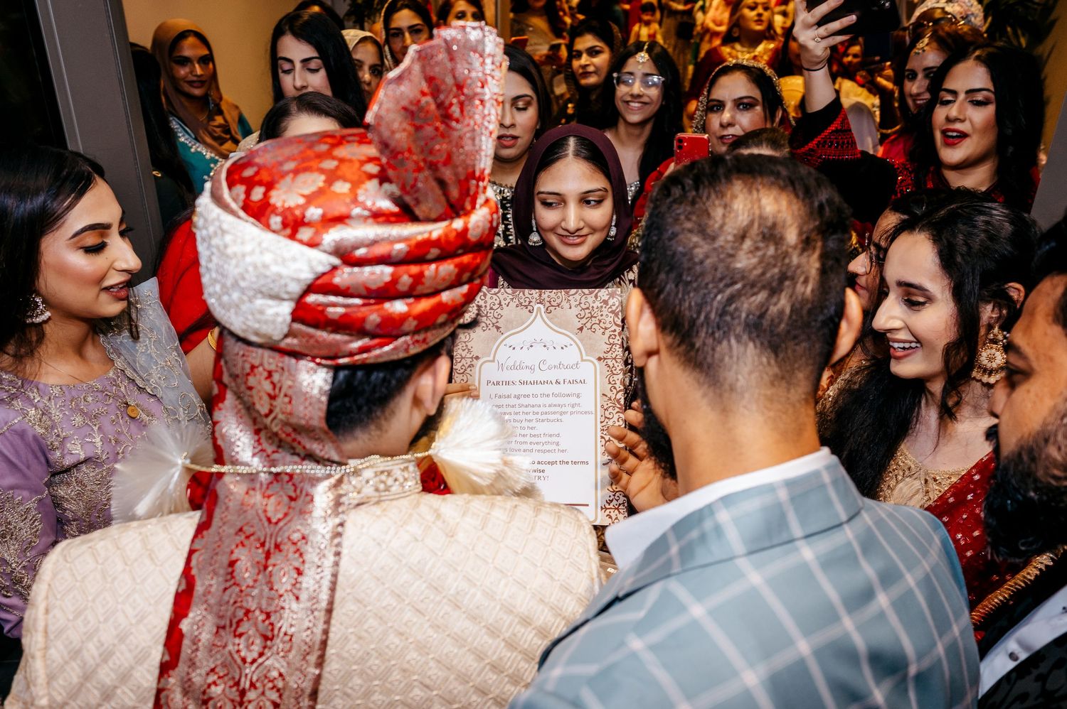 Indian groom in red sherwani and safa turban surrounded by guests at a vibrant traditional wedding celebration.