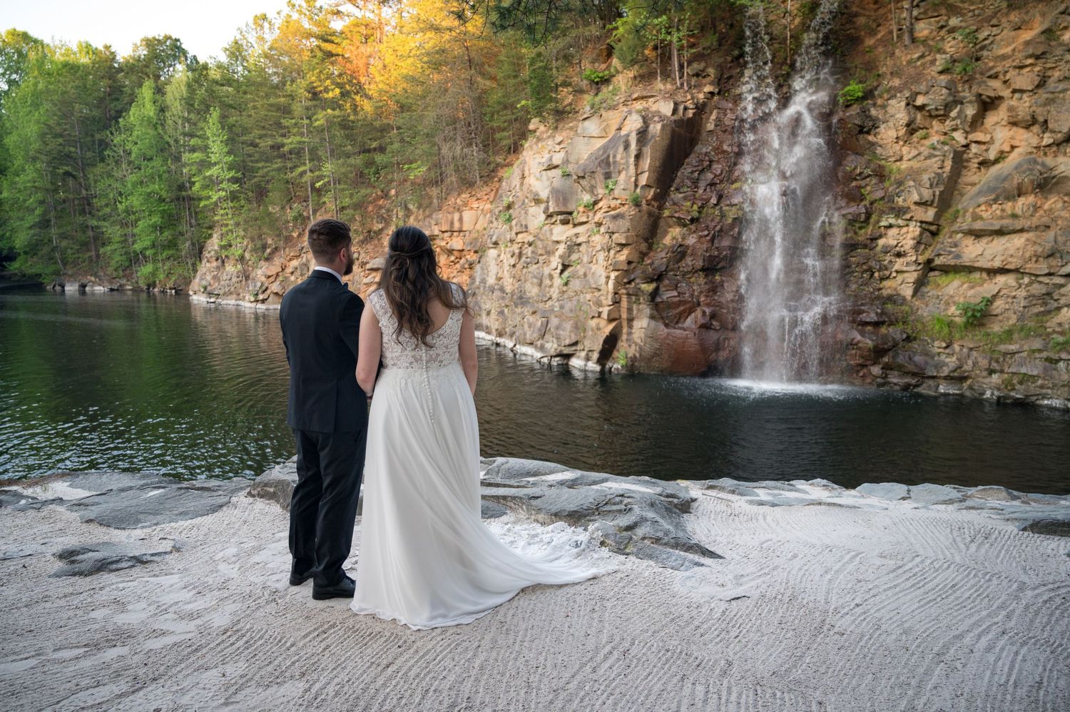 Bride and groom stand before a stunning waterfall and rocky cliffs at their outdoor wedding ceremony.