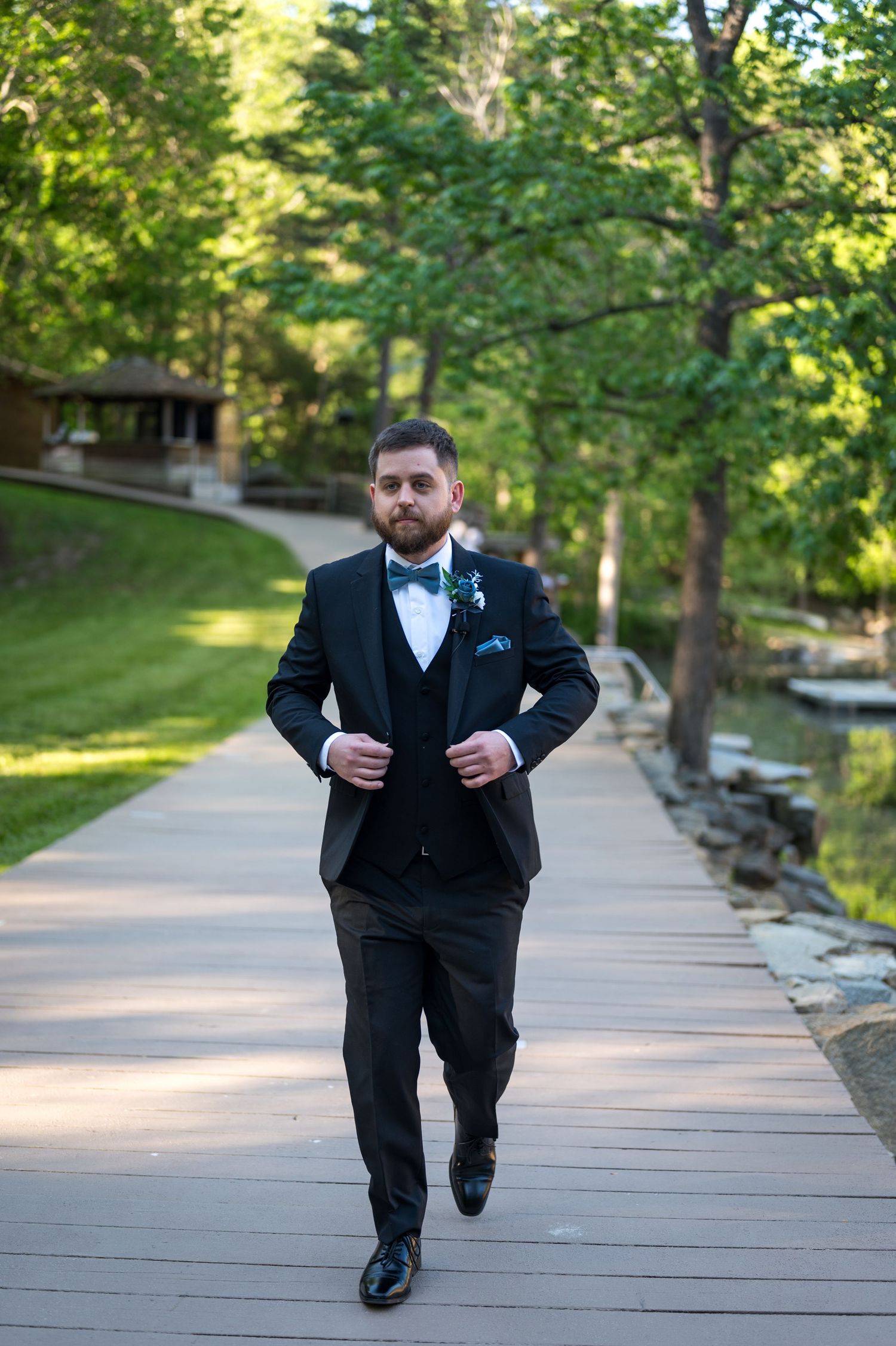 Groom in black tuxedo with bow tie and boutonniere walks confidently on wooden boardwalk surrounded by lush green trees.