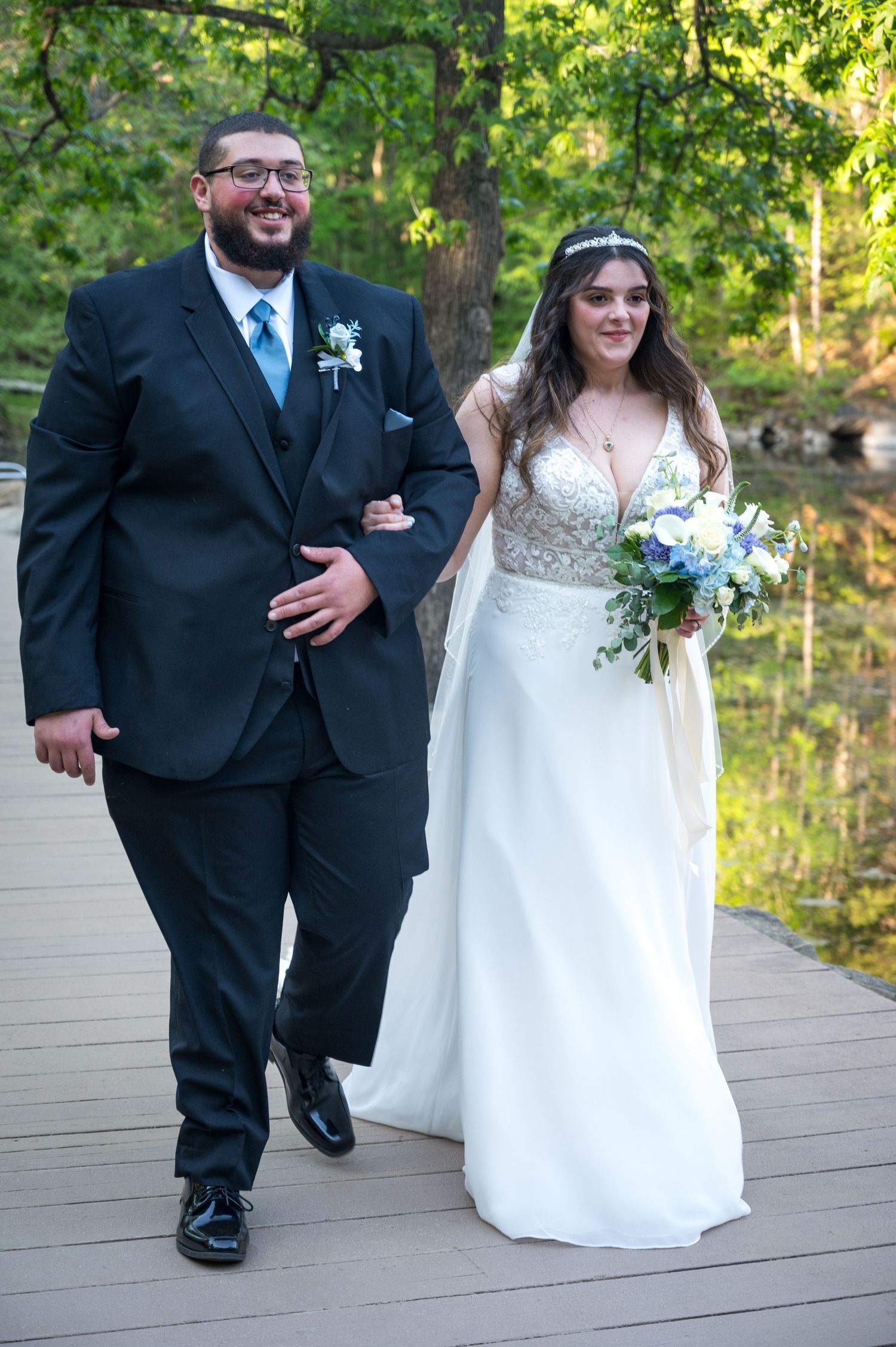 A smiling bride in a white gown holding blue flowers walks arm-in-arm with a groom in a black suit on a wooden deck.