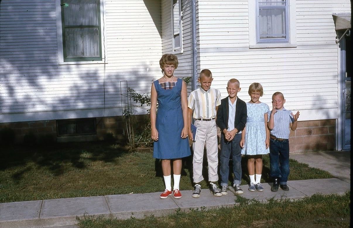 Five adults smiling together in front of a rustic shed surrounded by trees on a green lawn.