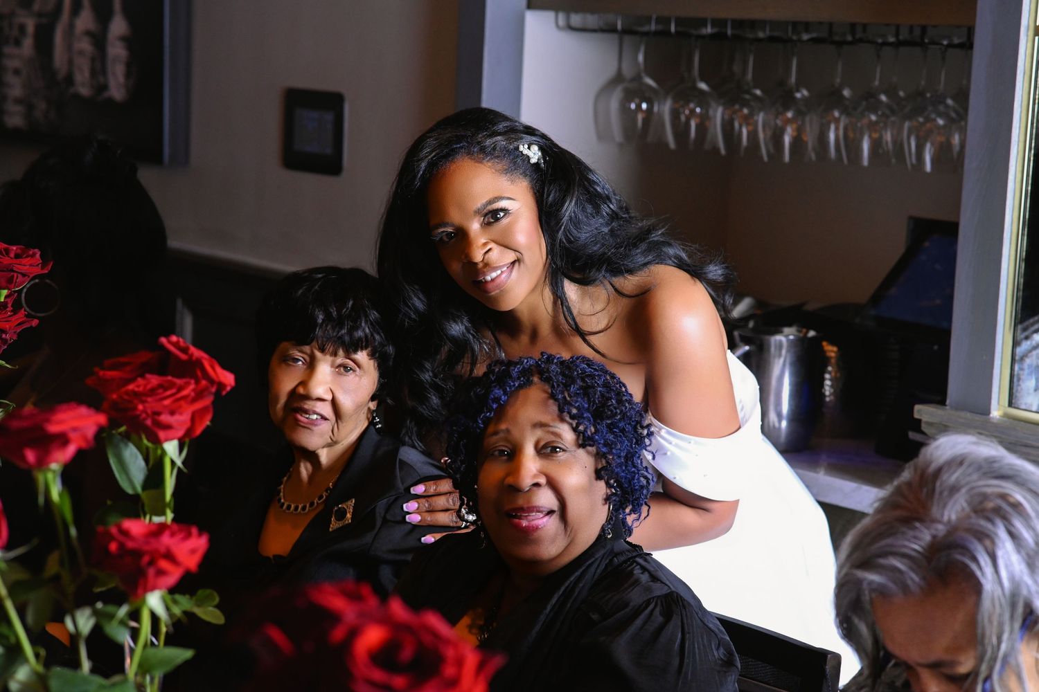 Three women smile together at a restaurant table adorned with red roses, with wine glasses hanging in the background.