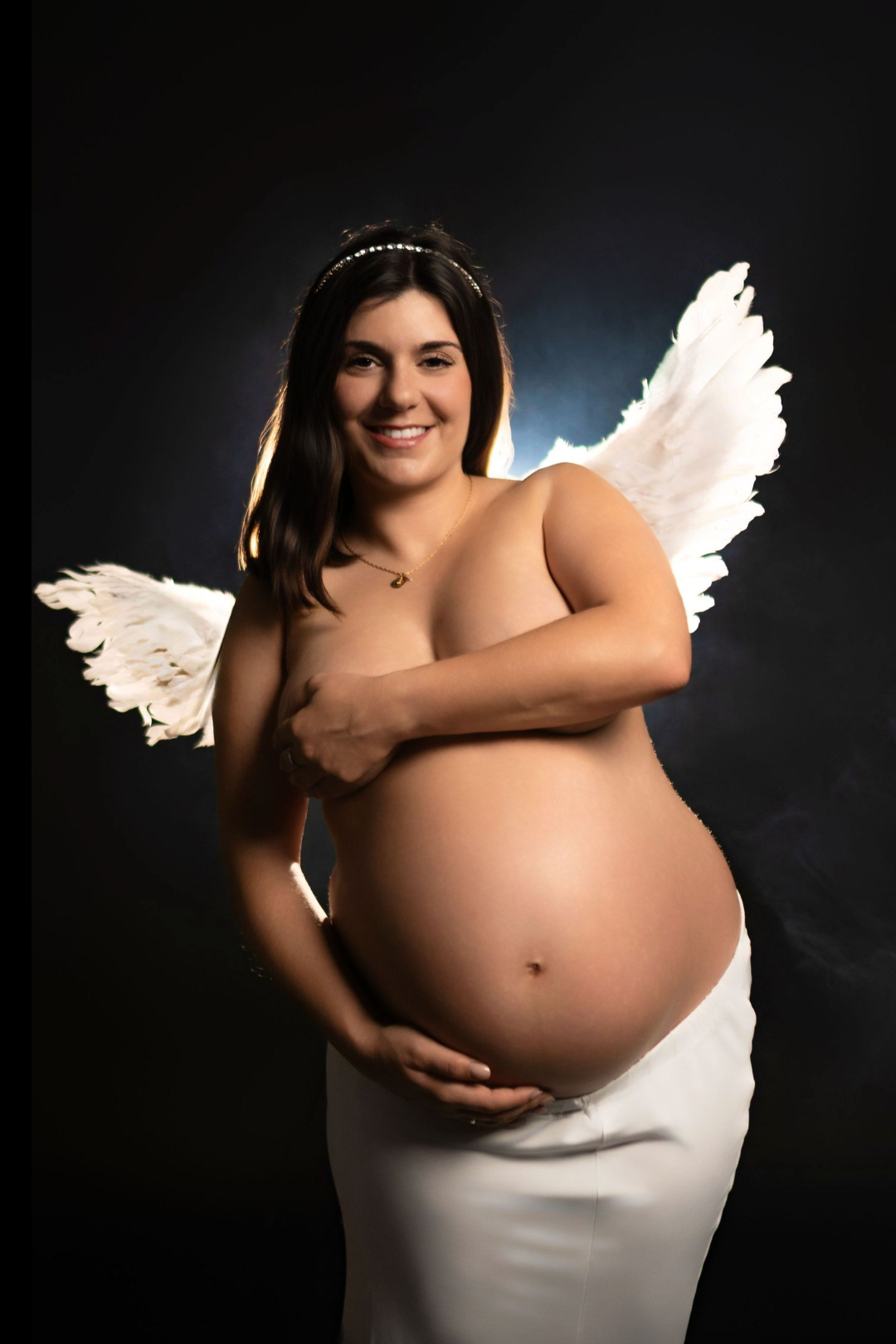 Smiling pregnant woman wearing angel wings and halo headband, cradling bare baby bump in white skirt on dark background.