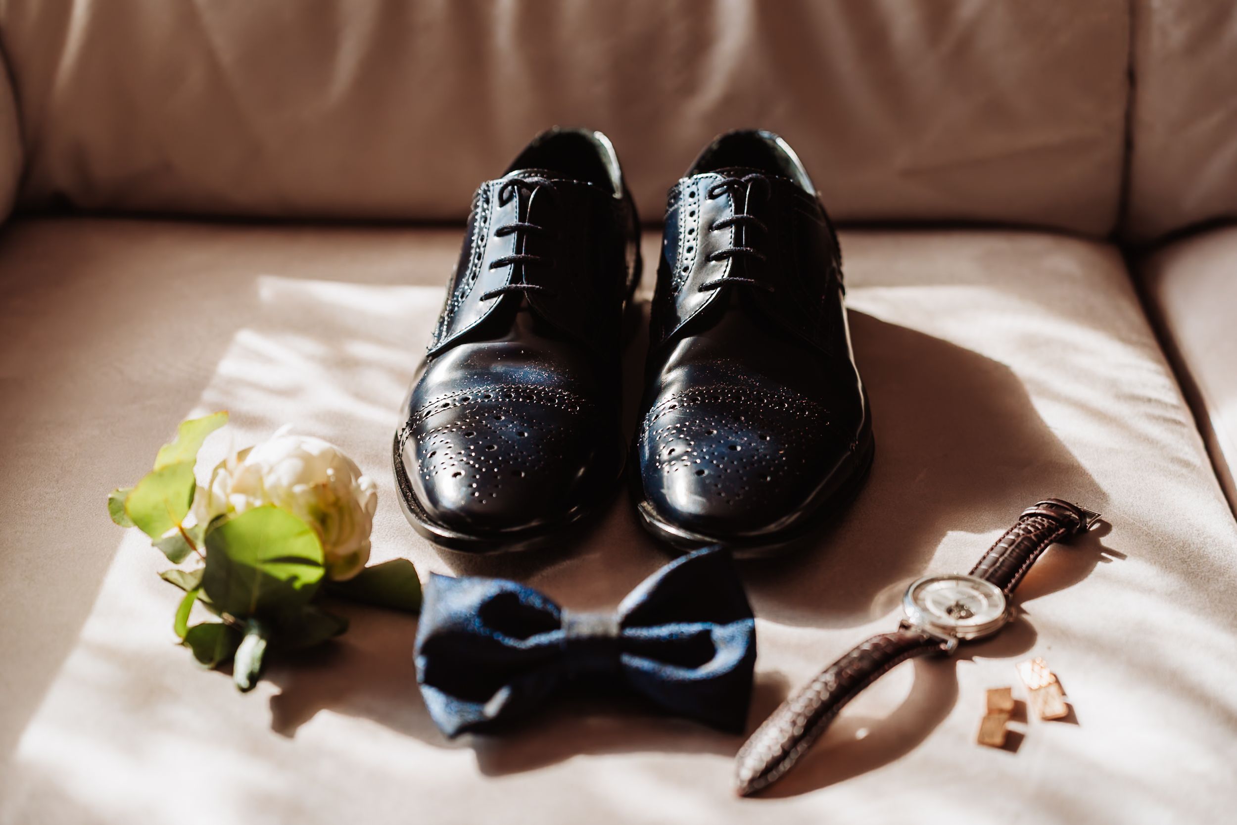 Black leather brogue dress shoes with bow tie, boutonniere, cufflinks, and watch arranged on a beige sofa.