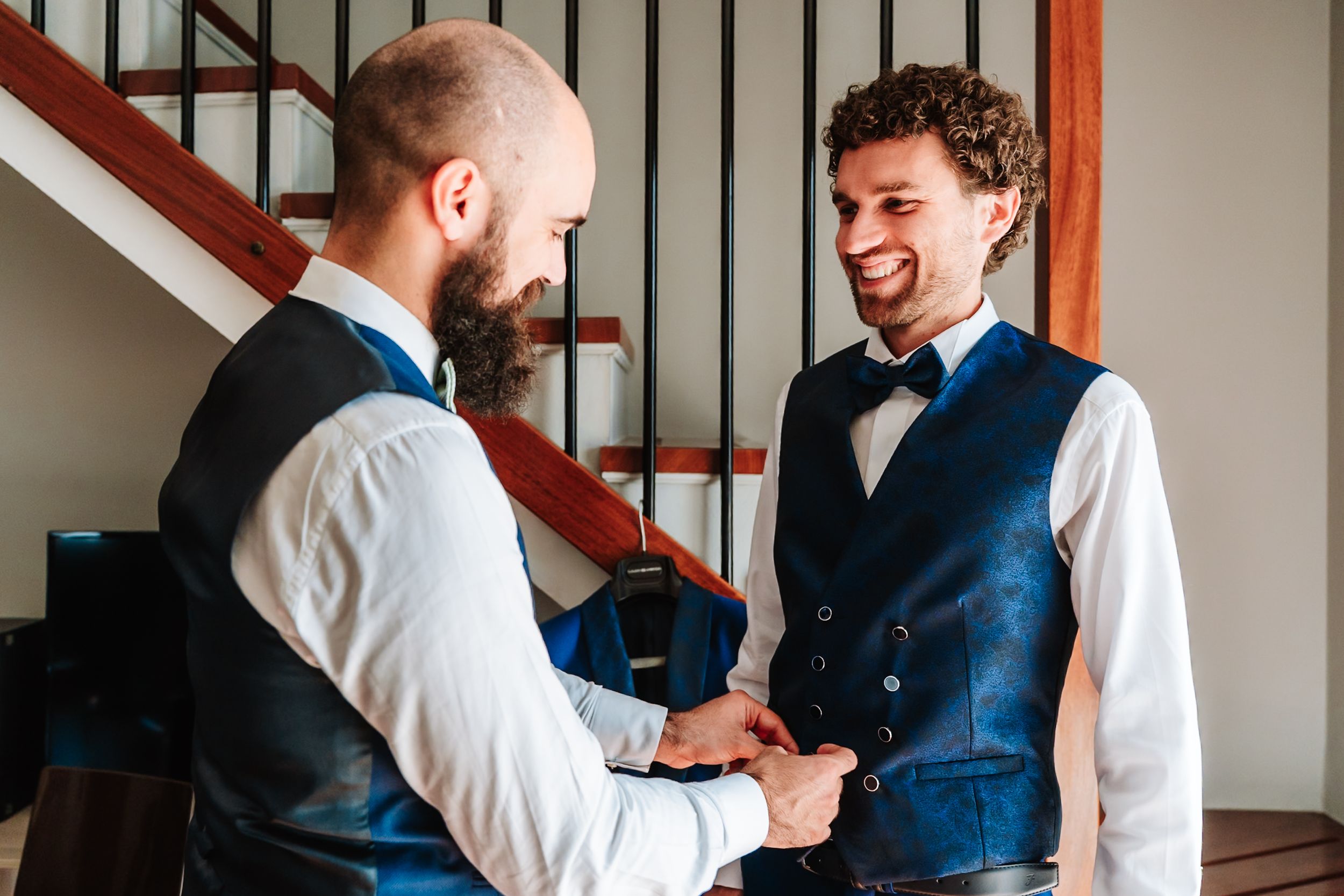 Two grooms in blue vests and bow ties smiling and holding hands during wedding preparations indoors.