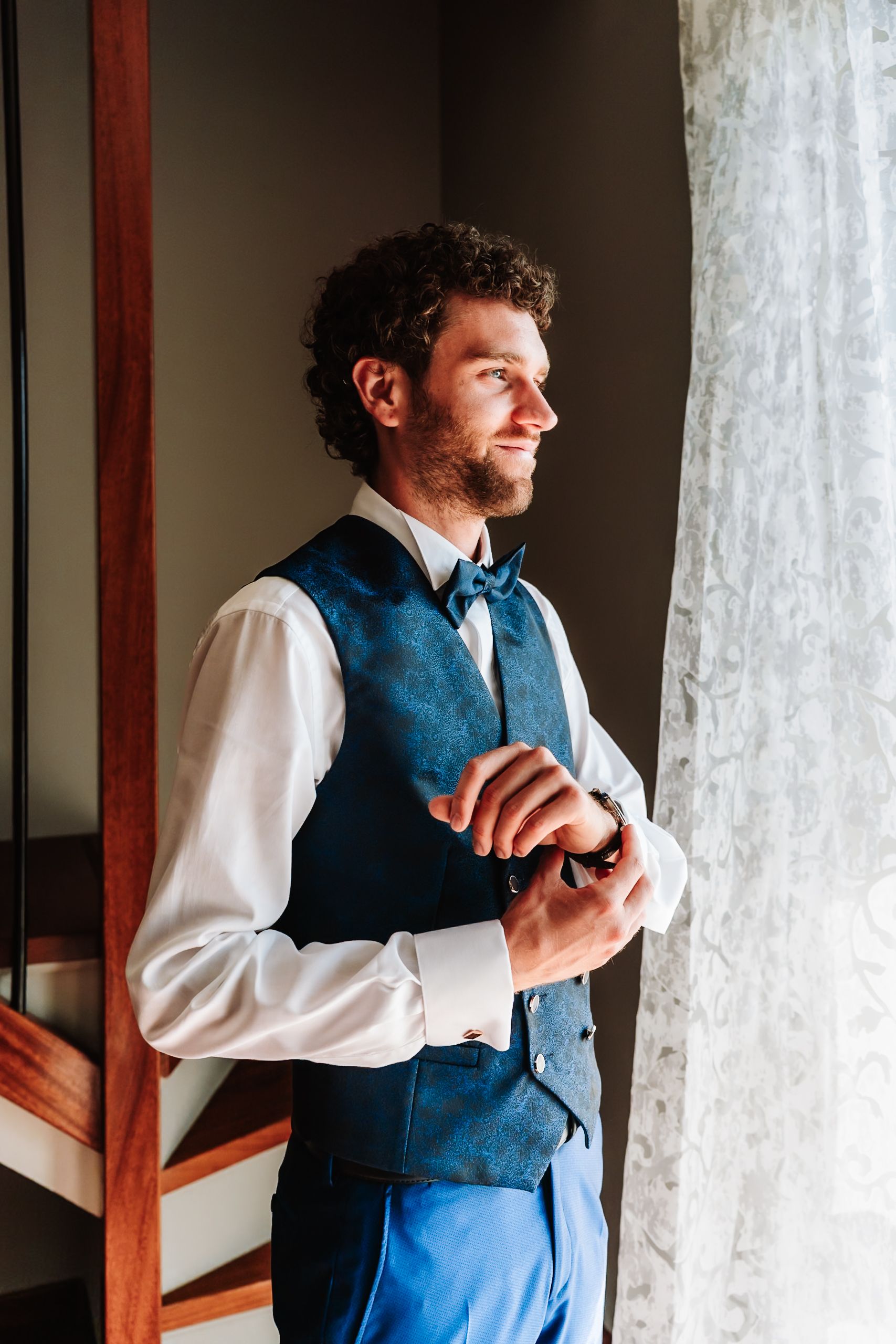 A well-dressed groom in a blue vest and bow tie adjusts his cufflinks by a sunlit window.