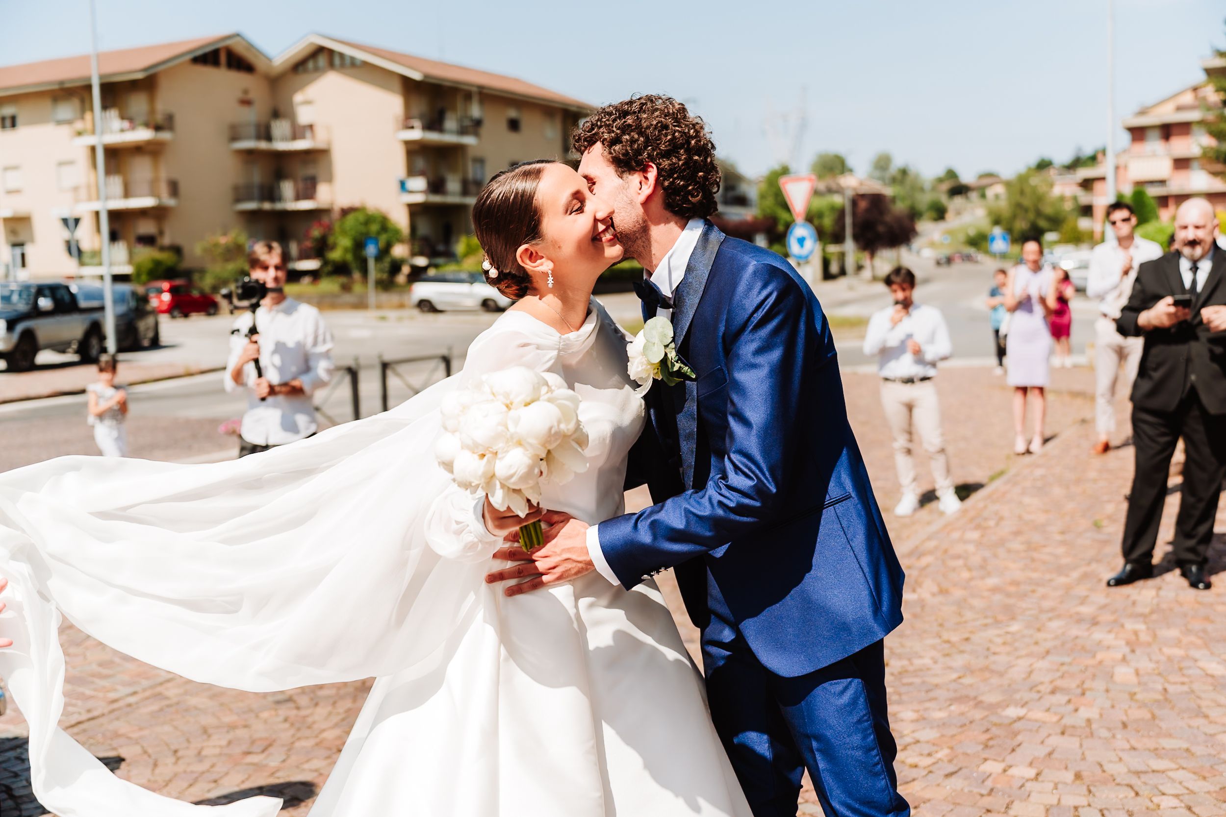 Bride and groom share a romantic kiss outdoors at their wedding, bride holding white bouquet with dress flowing in the breeze.