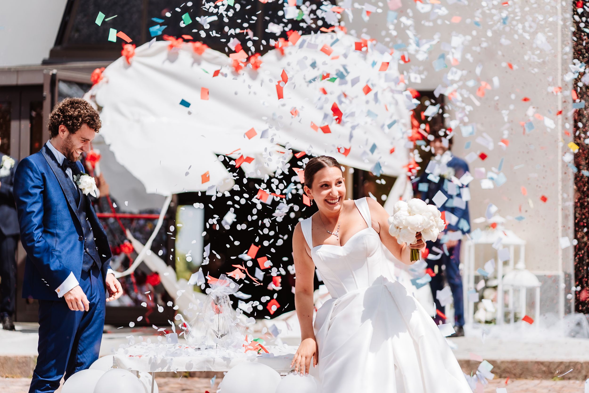Joyful newlyweds celebrate outdoors with colorful confetti, bride in white gown holding bouquet, groom in blue suit.