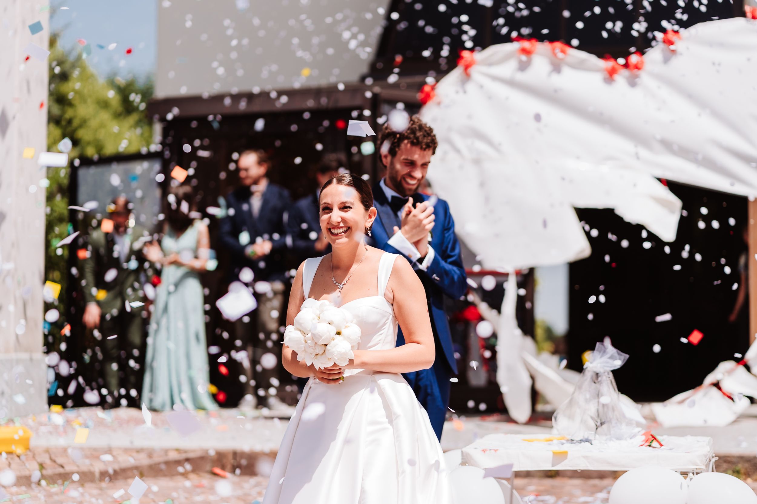Bride and groom walking through colorful confetti shower at outdoor wedding ceremony with guests celebrating in background.