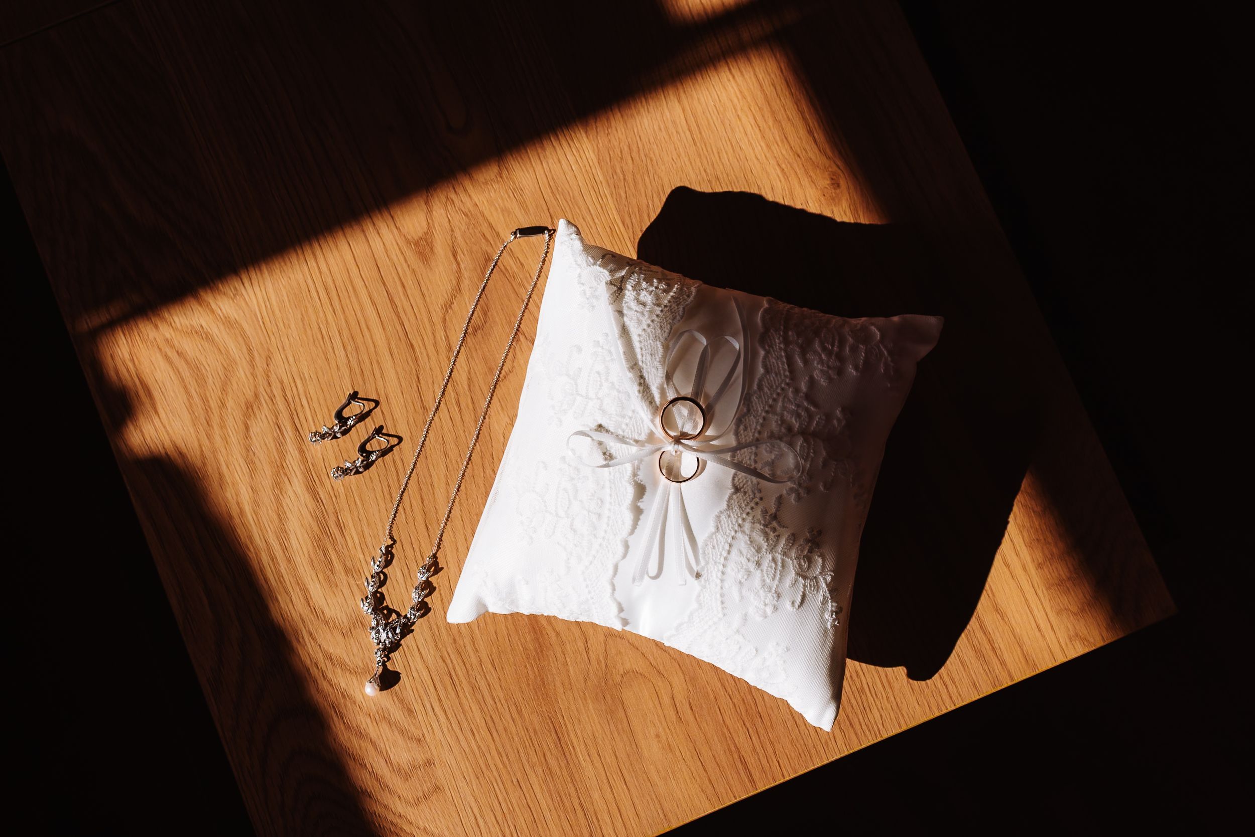 Wedding rings on white satin pillow with delicate necklace and earrings on wooden table in warm sunlight.