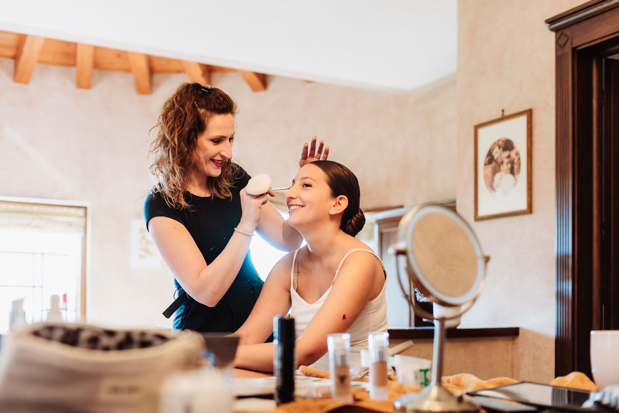 A makeup artist applying cosmetics to a smiling bride at a vanity mirror during wedding preparations.