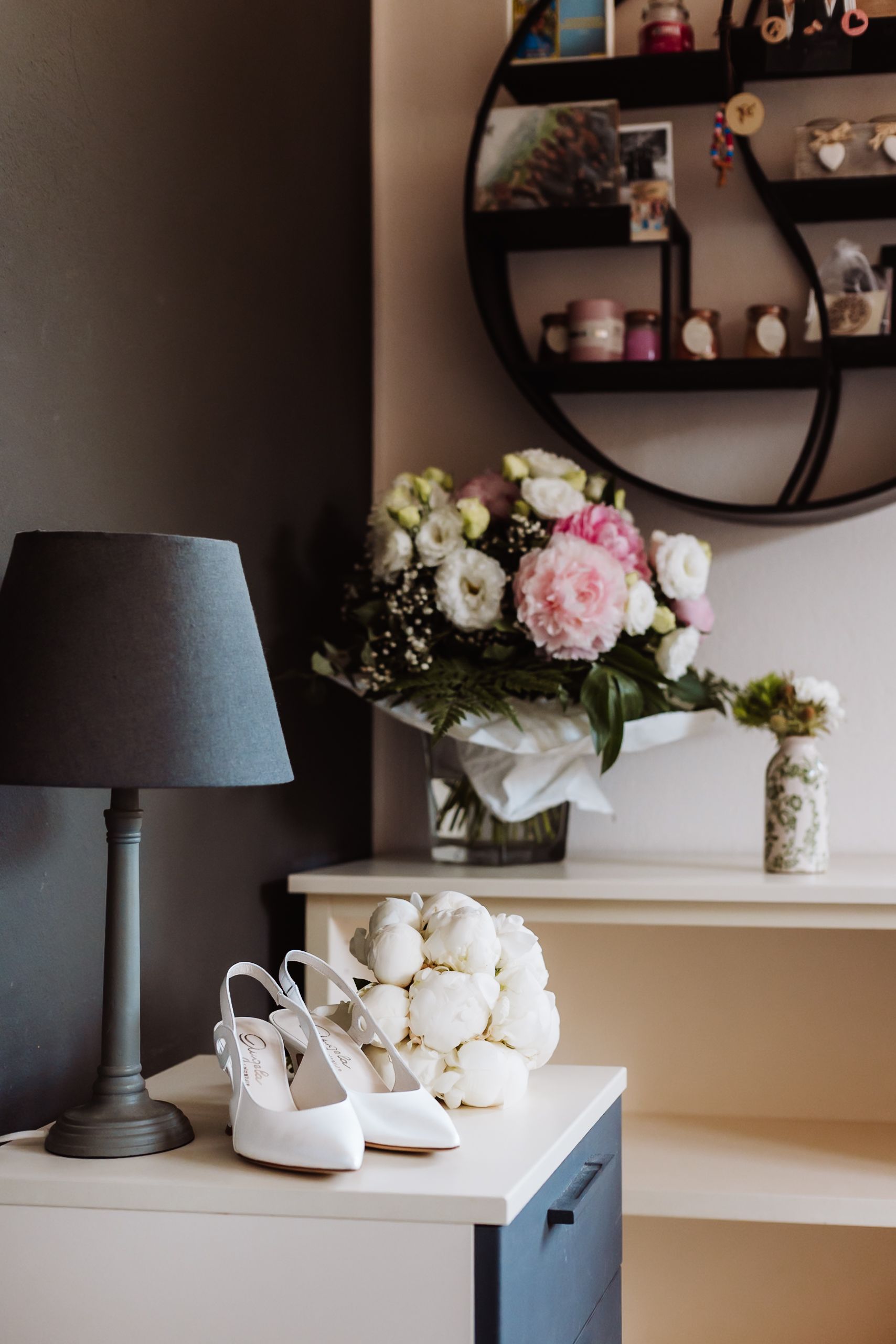 Elegant bridal setup with white heels, peony bouquet, lamp, and floral arrangement on a dresser with round mirror.
