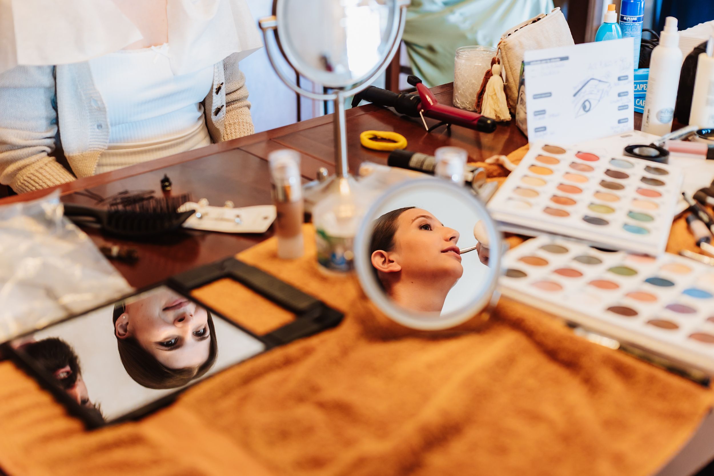 A makeup vanity table with mirrors reflecting a woman's face, surrounded by eyeshadow palettes and beauty products.