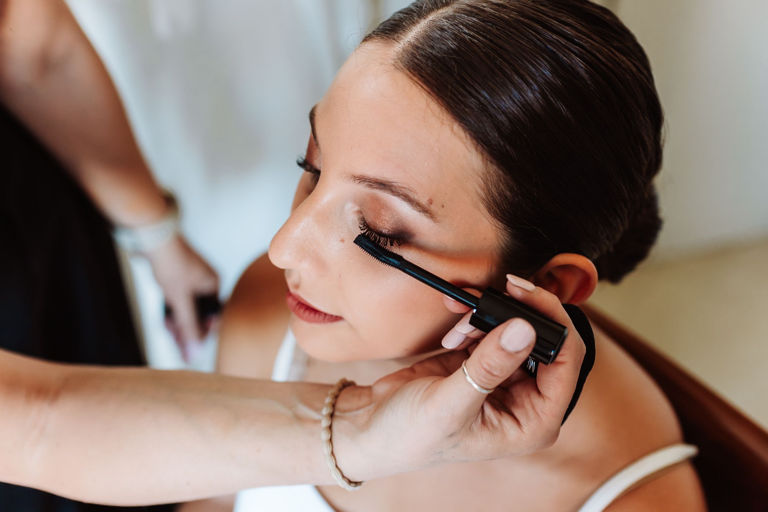 A makeup artist applies mascara to a bride with an elegant updo hairstyle before her wedding ceremony.