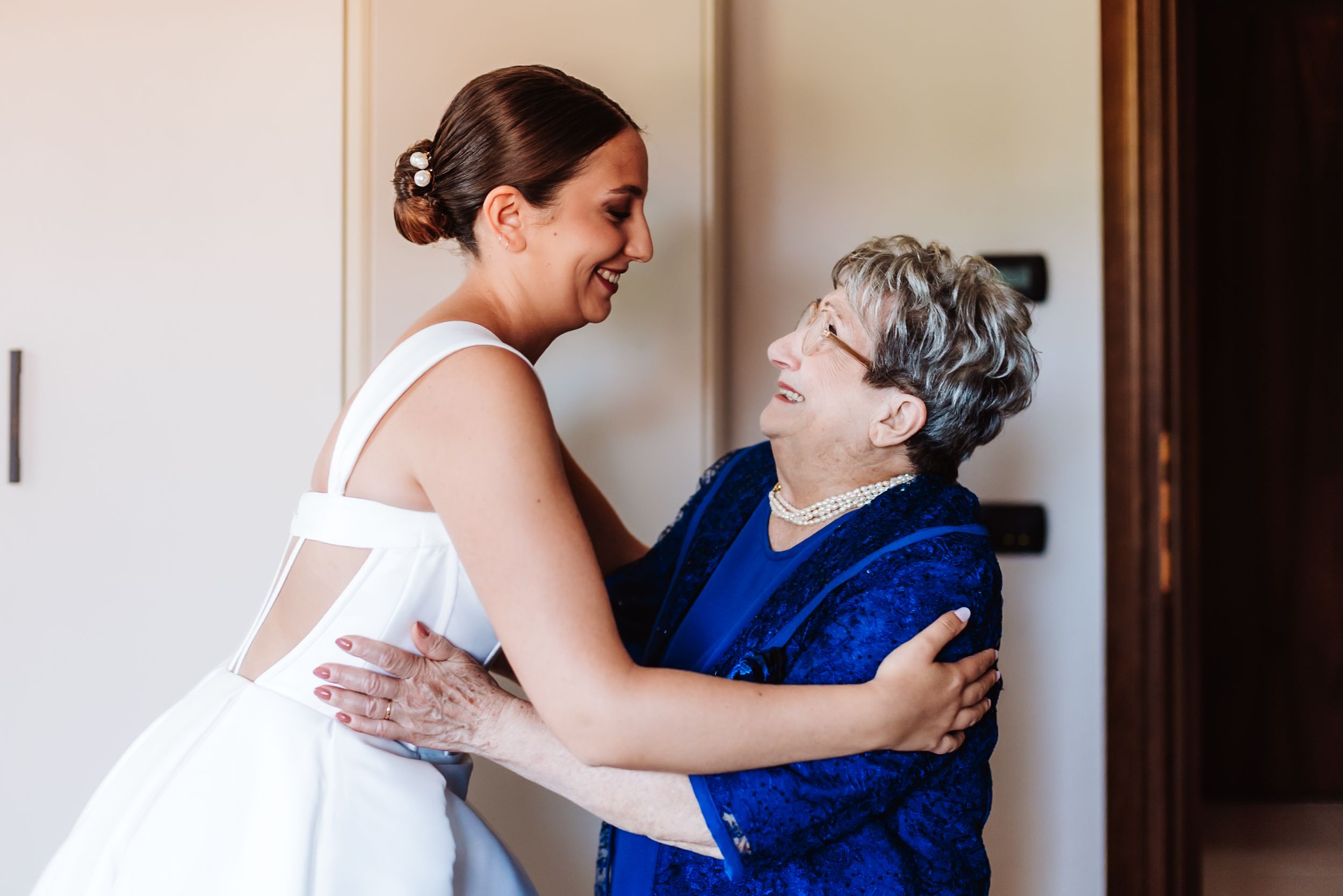 Bride in white gown embracing older woman in blue at wedding venue, sharing a joyful moment.