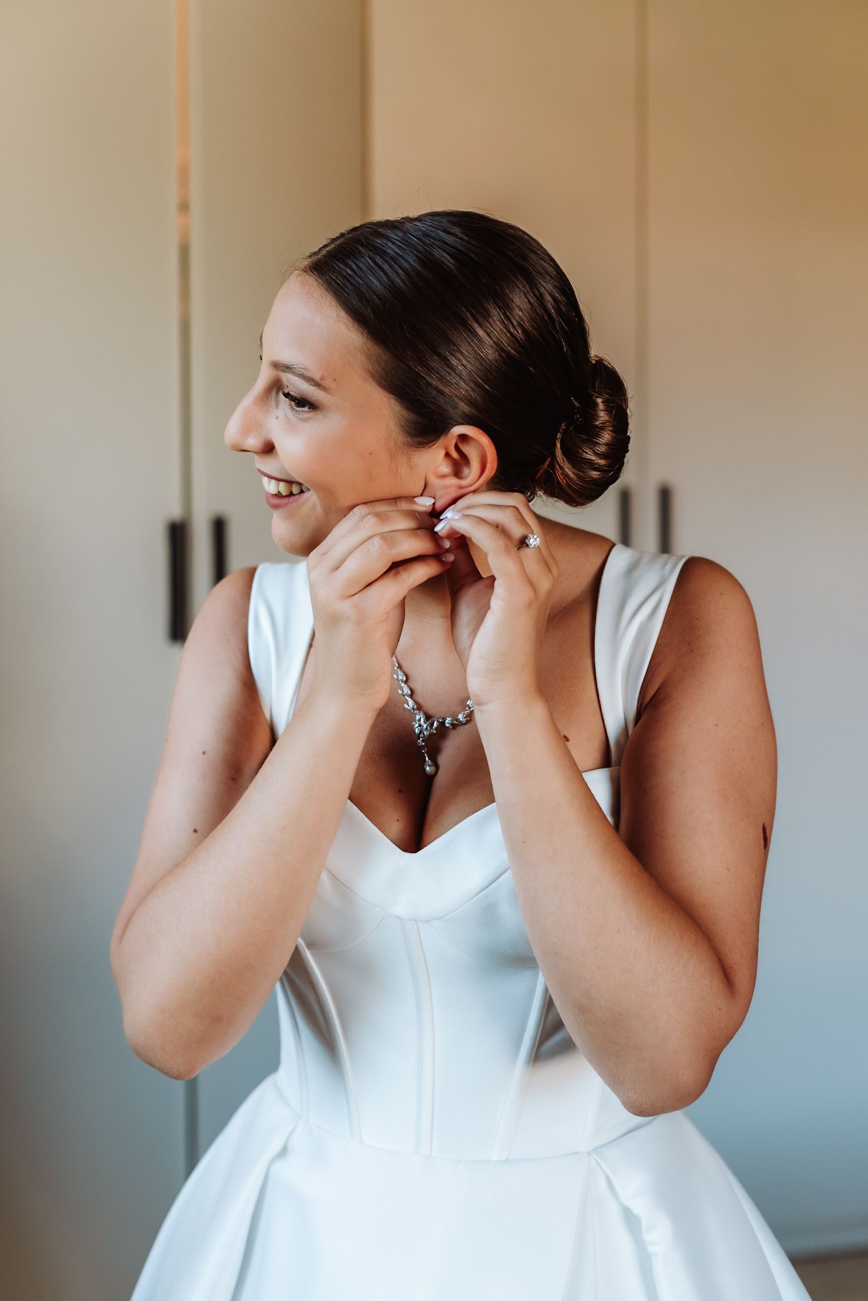 Bride in white gown putting on earrings, hair in elegant updo, wearing diamond bracelet and necklace.
