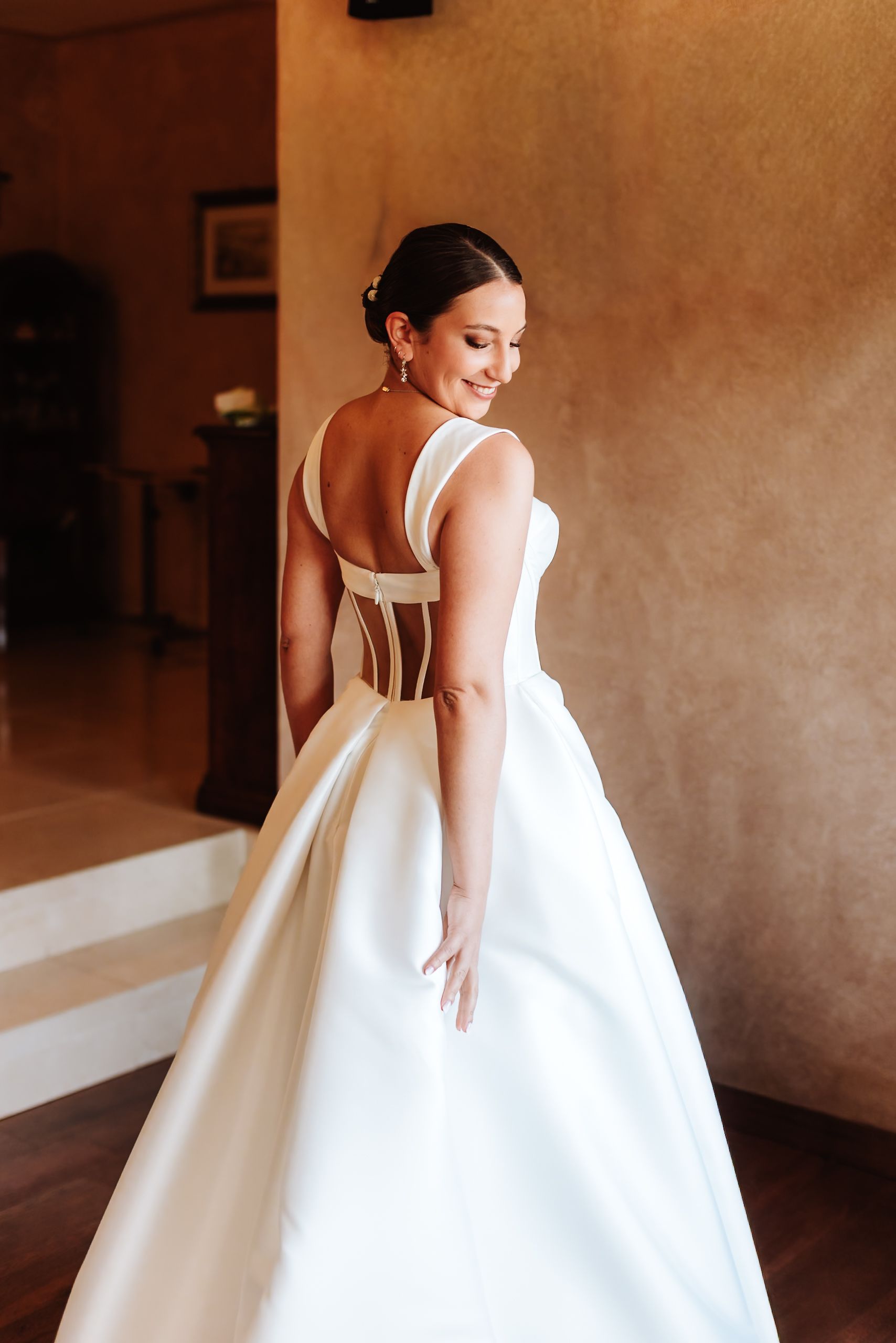Bride in elegant white backless wedding gown with corset detail, smiling over shoulder in warmly lit room.