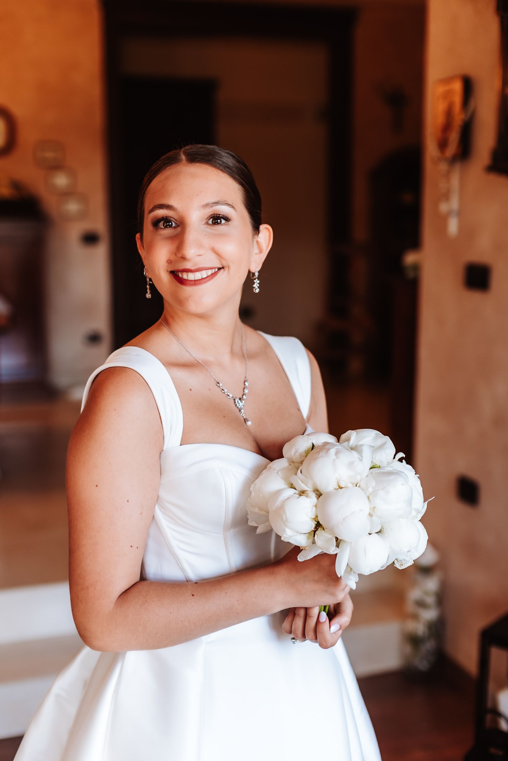 Smiling bride in white gown holding white peony bouquet, wearing diamond jewelry, standing indoors.