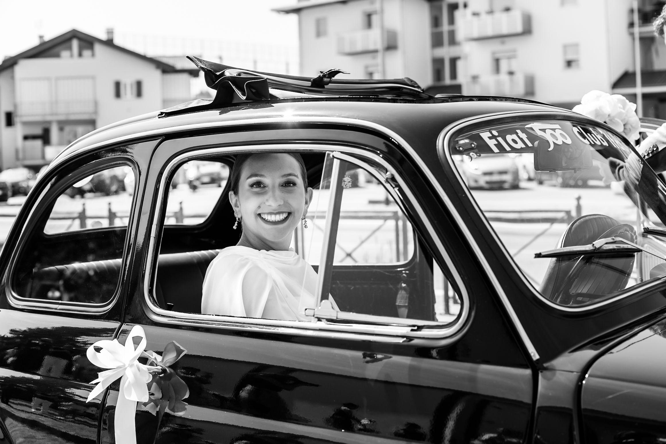 Smiling bride sitting in a vintage black Fiat car, black and white wedding photography.