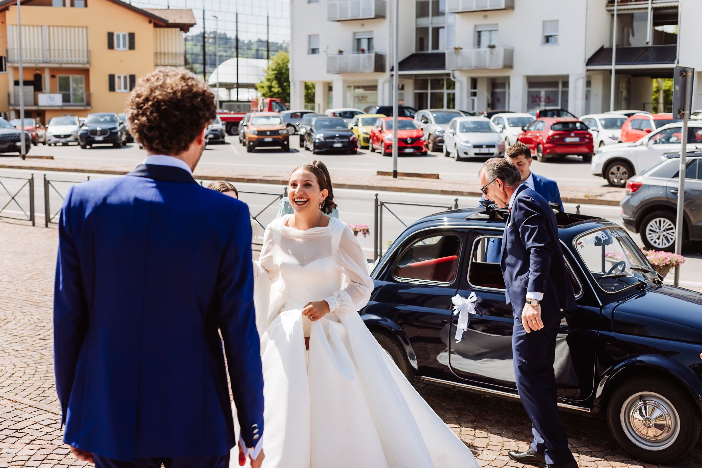 Bride in white gown arriving at wedding venue in classic black car, greeted by groom in navy suit.