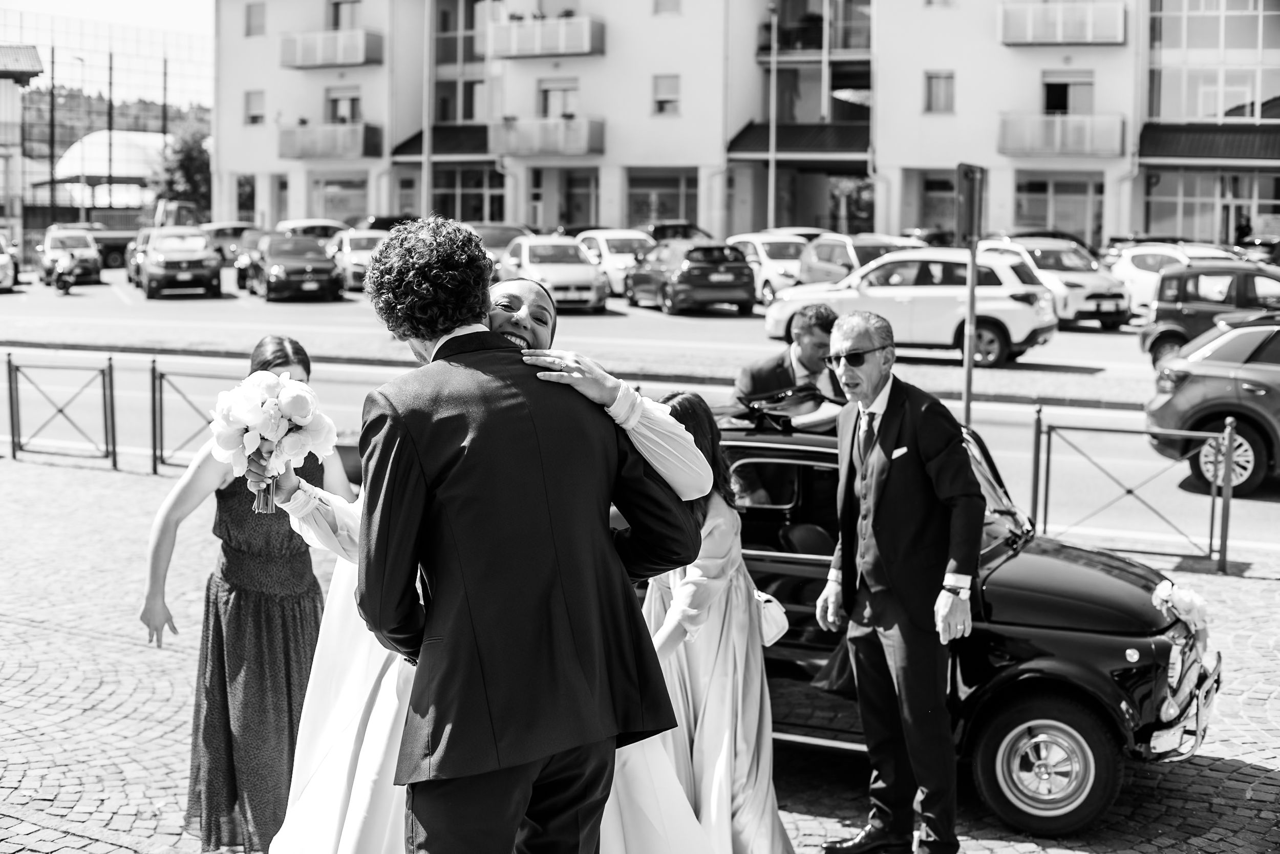 Black and white wedding photo of bride and groom arriving at venue with classic car and guests outside.